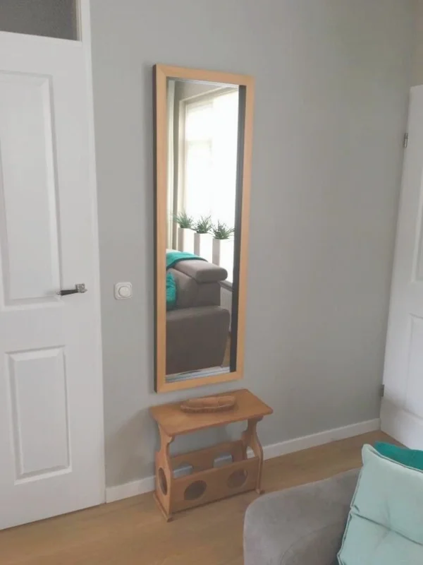 Deze foto laat een rustige hoek zien uit de woonkamer & eetkamer make-over, met een staande spiegel in een warme houten lijst en een compact houten bijzetmeubel. De lichte wand en natuurlijke houttinten zorgen voor een frisse, open uitstraling, terwi