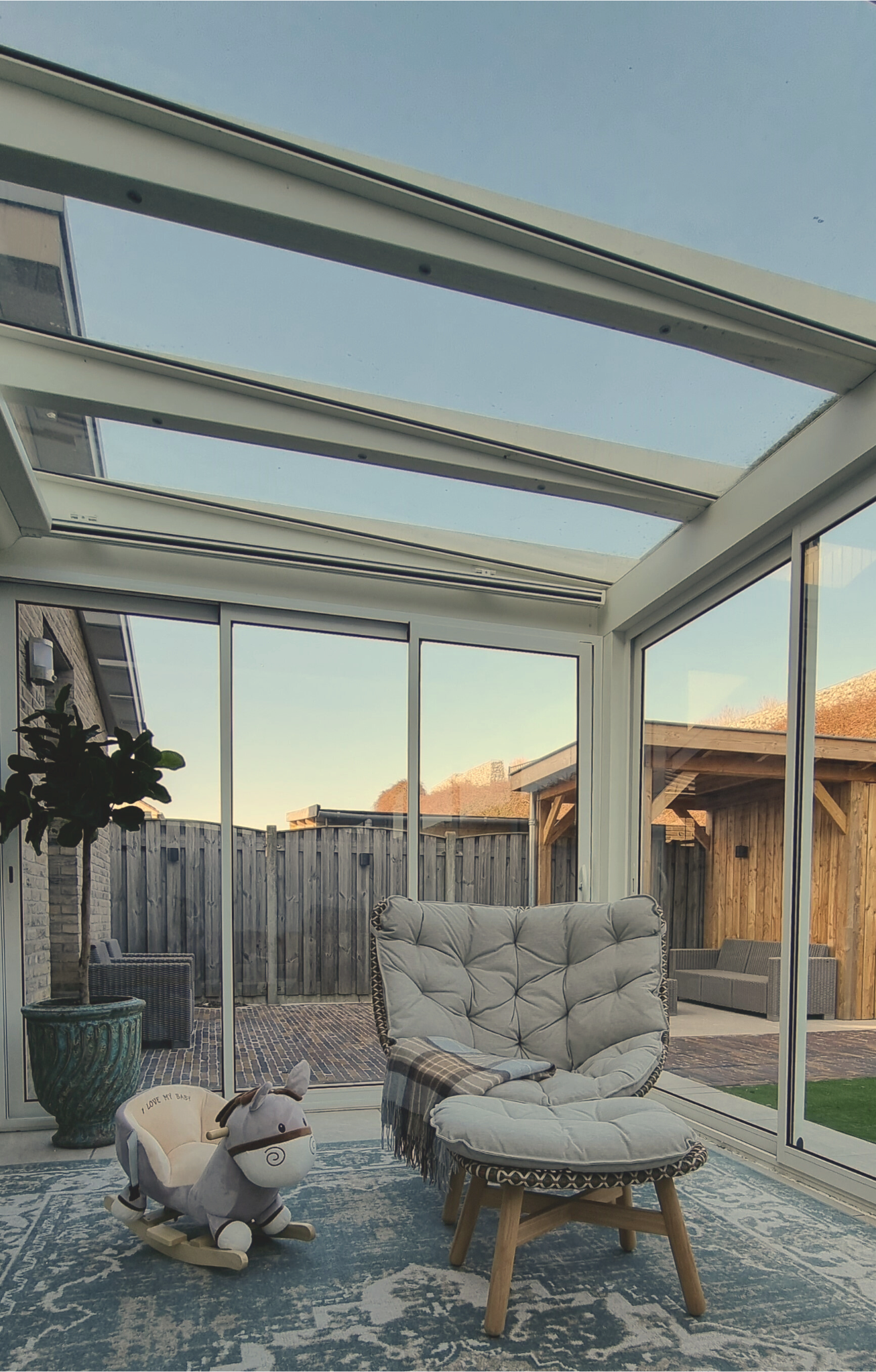 Lichte veranda met glazen dak en glazen wanden, ingericht met een relaxstoel, voetenbank en vloerkleed.