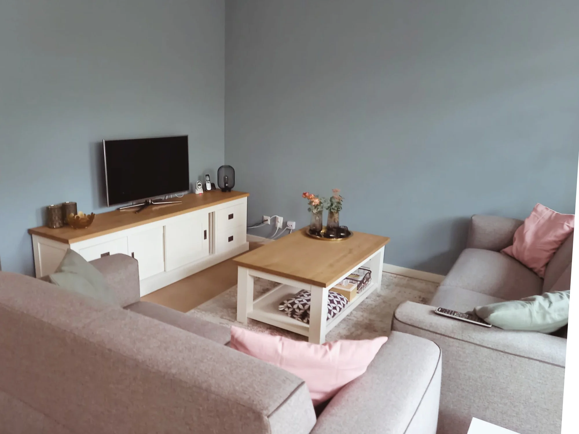 Rustige woonkamer met grijze hoekbank, zachte roze accenten en licht houten meubels. Neutrale basis met warme styling voor een moderne, comfortabele en casa — overzichtelijk, sfeervol en in balans.  Interieuradvies, interieurontwerp en styling door E