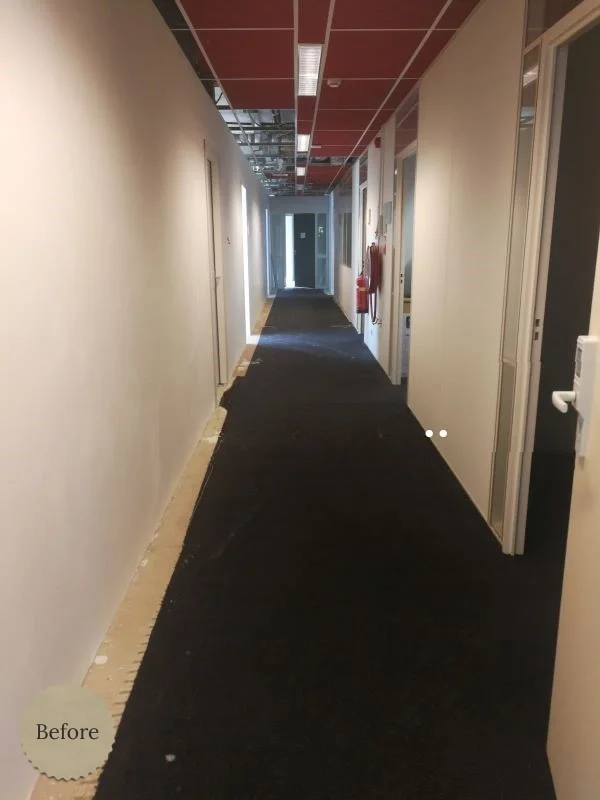 Voor de verbouwing: lange kantoor-/bedrijfsgang met witte wanden, donkere tijdelijke vloerbedekking en zichtbare installaties aan het plafond. Ideale “before” foto voor een renovatieproject van een moderne, warmere werkomgeving.
