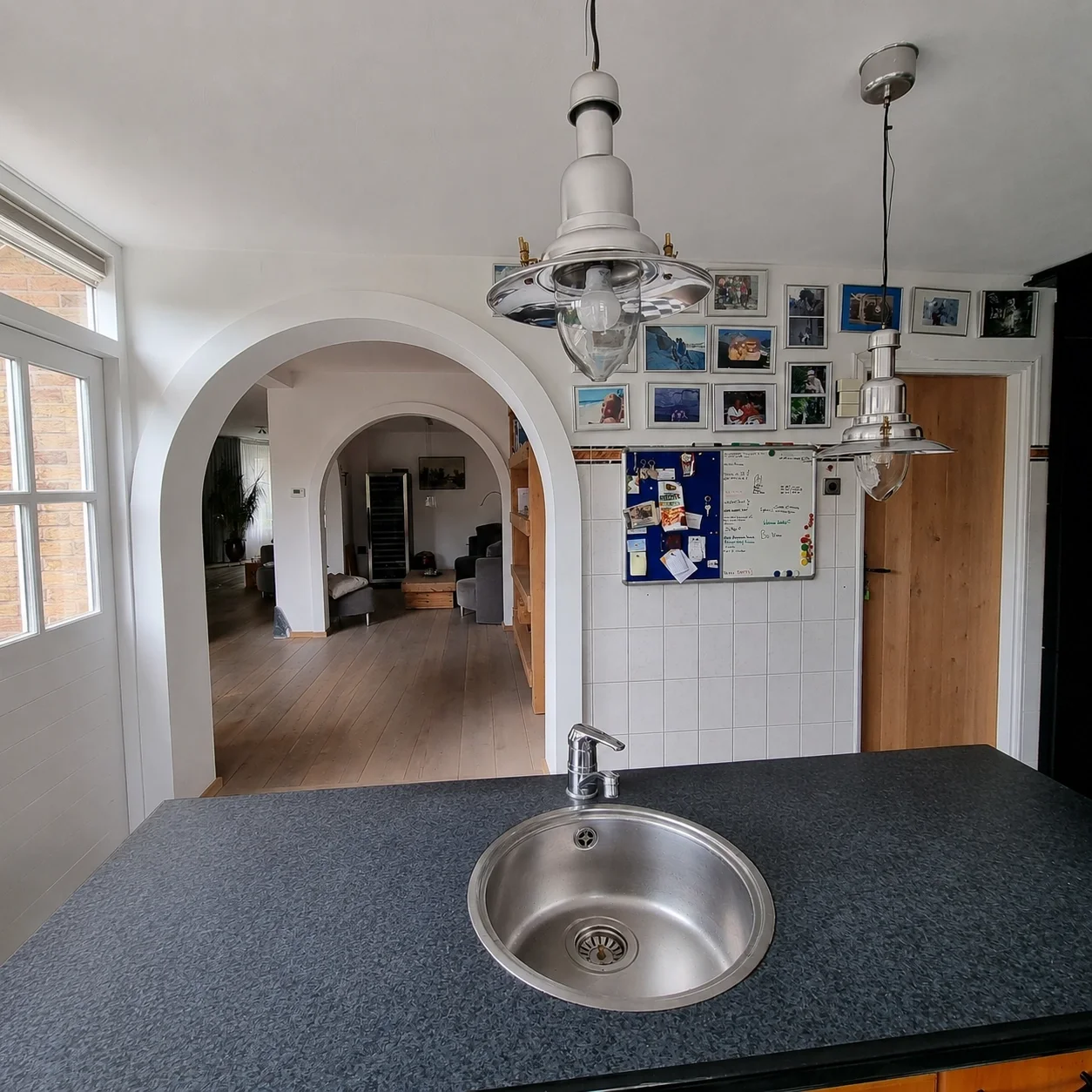 Before foto met zicht vanuit de keuken naar de woonkamer door een ronde boogdoorgang, met donker werkblad, spoelbak en hanglampen. Witte wanden en persoonlijke decoratie zorgen voor een huiselijke basis, maar de ruimte mist nog rust, eenheid en een m