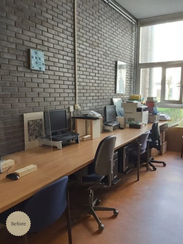 Before situatie van de kantoorinrichting met eenvoudige werkplekken langs een bakstenen muur, losse bureaus en beperkte opbergruimte, met daglicht vanuit het raam aan het einde van de ruimte.
