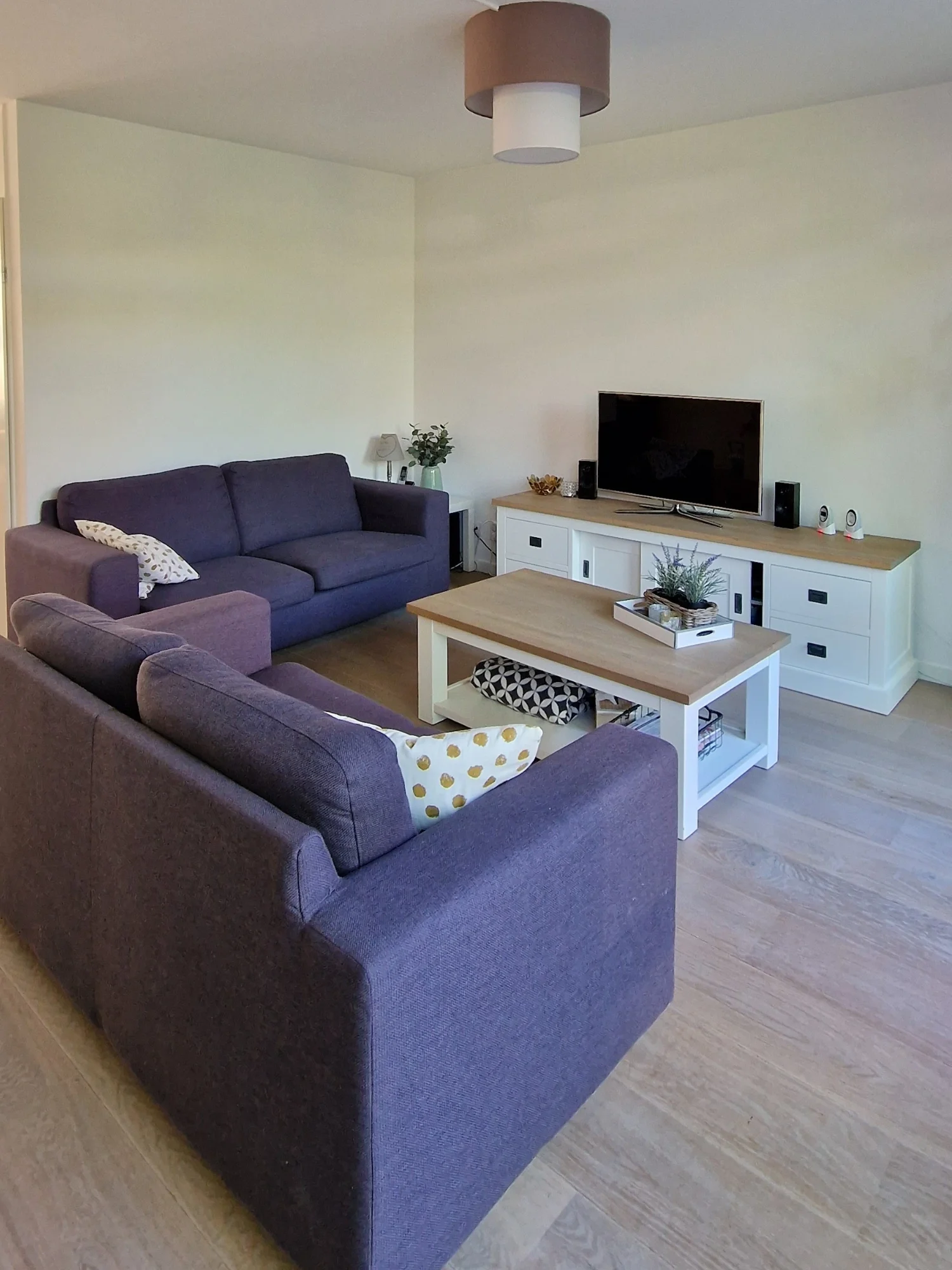Voorfoto van een compacte woonkamer met grijze hoekbank, witte salontafel en tv-meubel, met houtlook vloer en rustige basis.