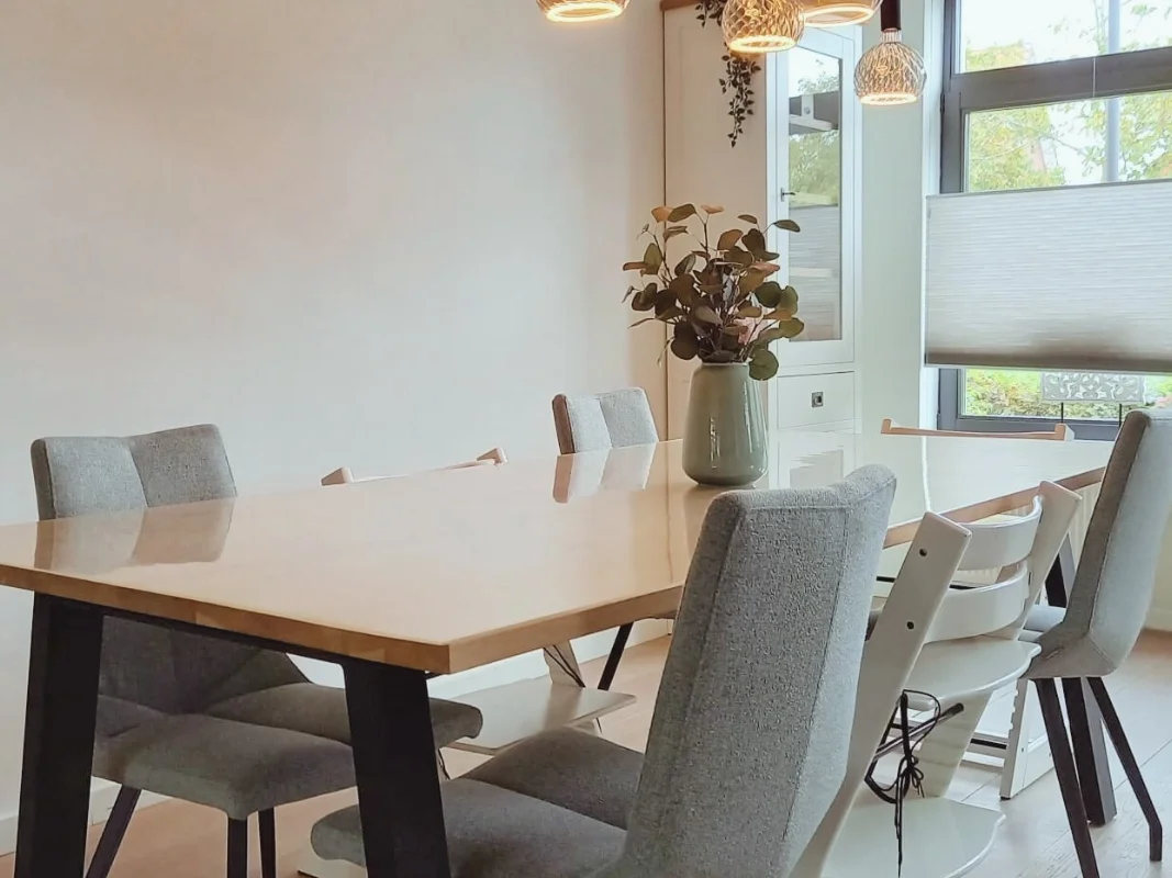 Moderne eethoek met houten eettafel, zachte stoffen stoelen en natuurlijke tinten. Een rustige, warme eetkamer met veel daglicht, passend bij een en casa interieur waar comfort en sfeer samenkomen.  Interieuradvies, interieurontwerp en styling door E