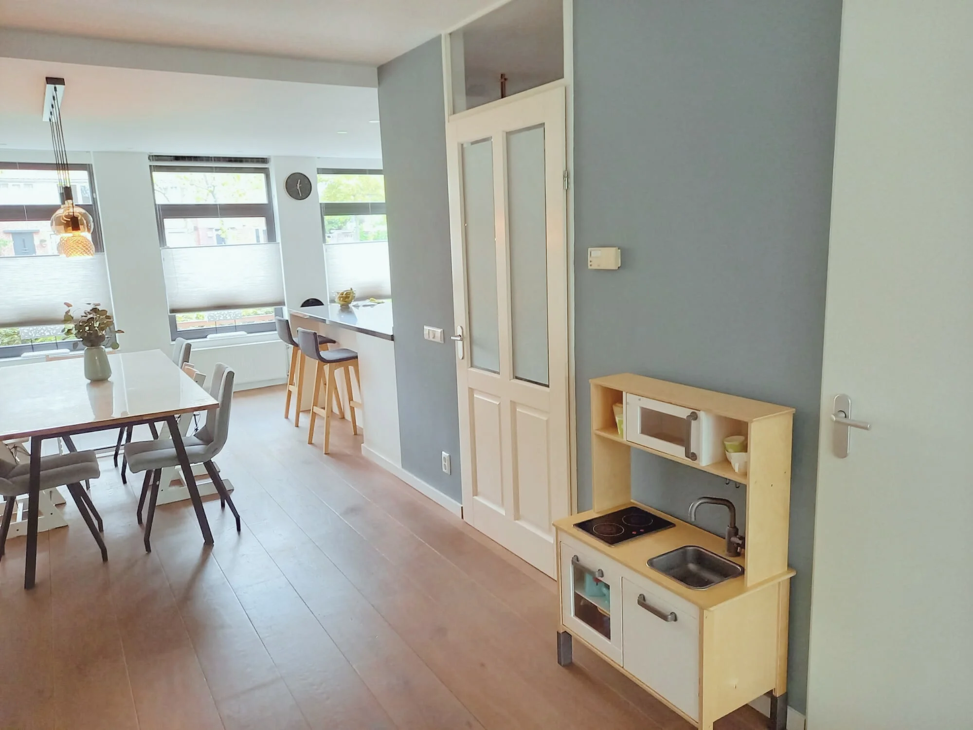 Open keuken en eethoek met veel daglicht, houten vloer en rustige blauwgrijze wand. Praktisch familie-interieur met natuurlijke materialen en een speelhoekje, waardoor wonen, eten en spelen mooi samenkomen — en casa in balans.  Interieuradvies, inter