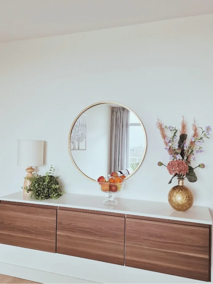 Wandmeubel met houten fronten en ronde spiegel als eyecatcher. Met bloemen, groen en een fruitschaal voor een warme, rustige styling.  Interieuradvies, interieurontwerp en styling door En-Casa Interiors in Noord-Holland.