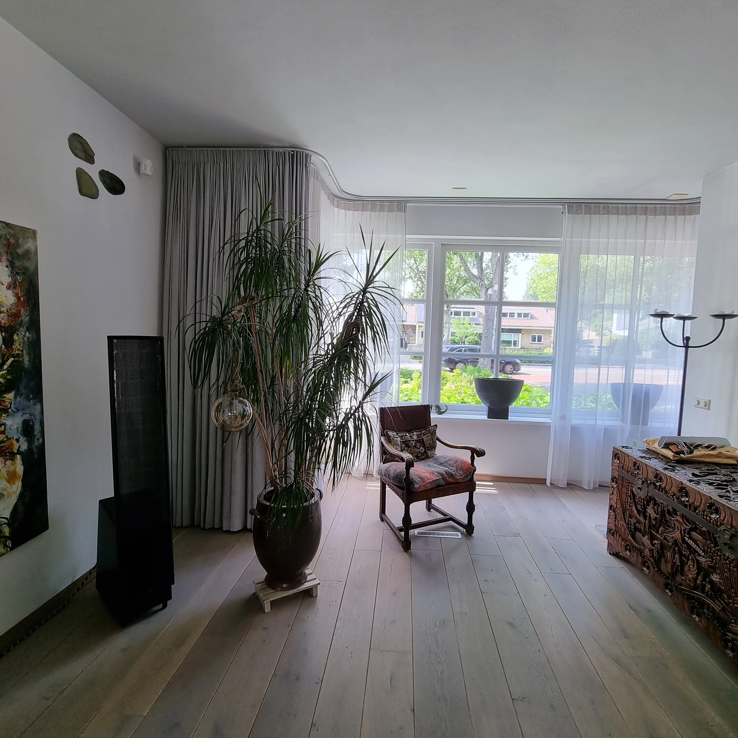 Deze foto toont een lichte voor-situatie van een woonkamer met grote raampartijen, een houten vloer en een rustige basis. De aanwezige meubels, planten en raambekleding geven inzicht in de bestaande indeling en vormen het vertrekpunt voor een nieuw i