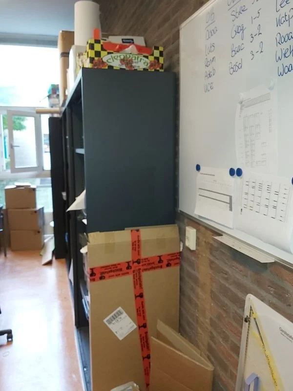 Before situatie van het kantoor met rommelige opslag en zichtbare stapels dozen door gebrek aan voldoende opbergruimte, waardoor de werkruimte onrustig en minder efficiënt aanvoelt.