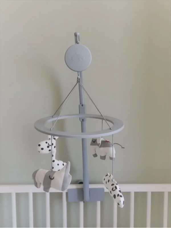 Close-up van een baby mobiel boven een wit ledikant. De mobiel is uitgevoerd in zachte grijs- en wittinten en heeft hangende figuurtjes, waaronder een olifantje en dieren met stippen. Tegen de rustige, zachtgroene muur zorgt dit detail voor een lieve