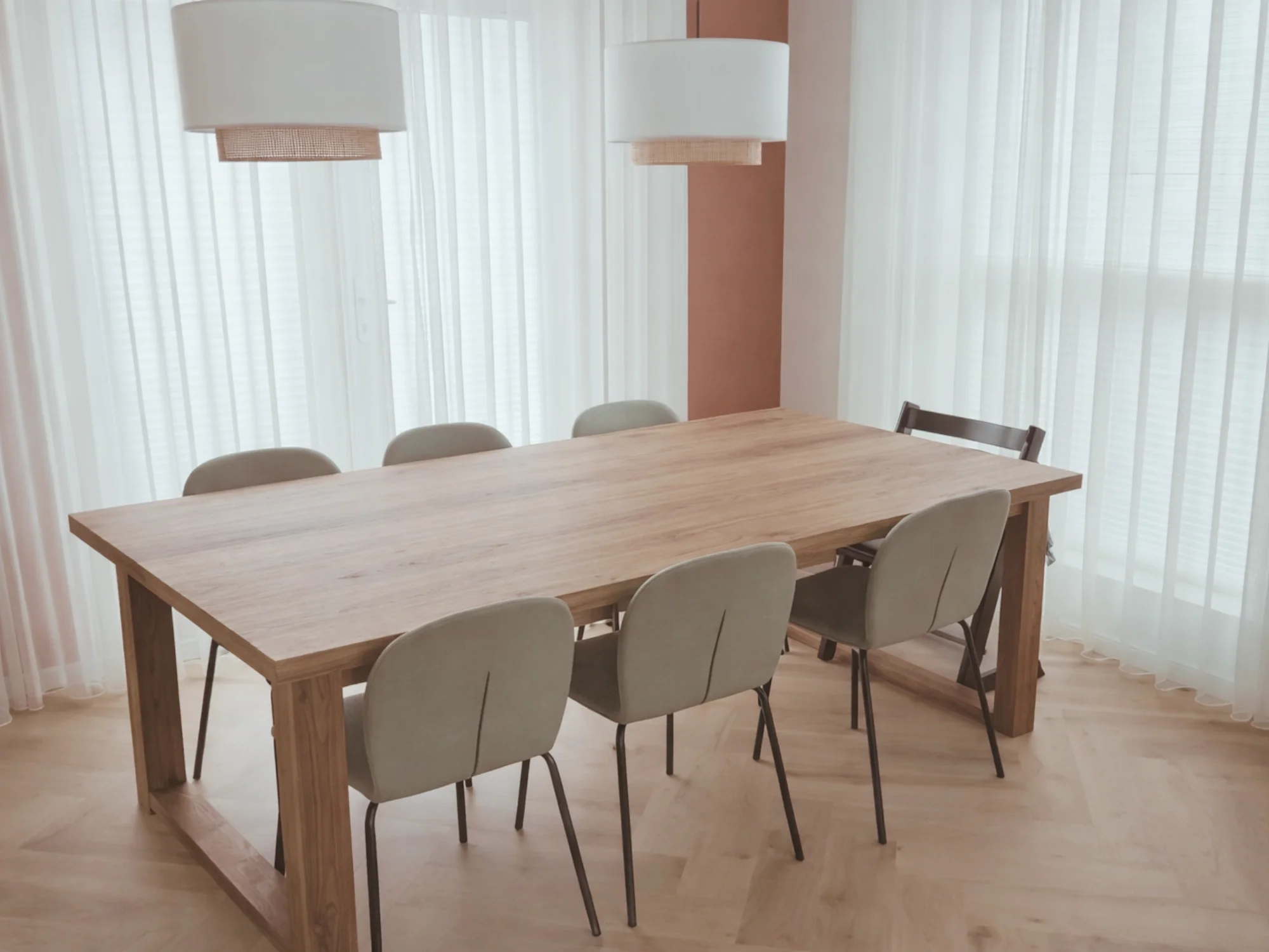 Eetkamer met een grote houten eettafel en zachte, beige eetkamerstoelen met zwarte poten. Boven de tafel hangen twee witte hanglampen met natuurlijke details. Grote ramen met transparante gordijnen zorgen voor veel daglicht en een warme, rustige sfe