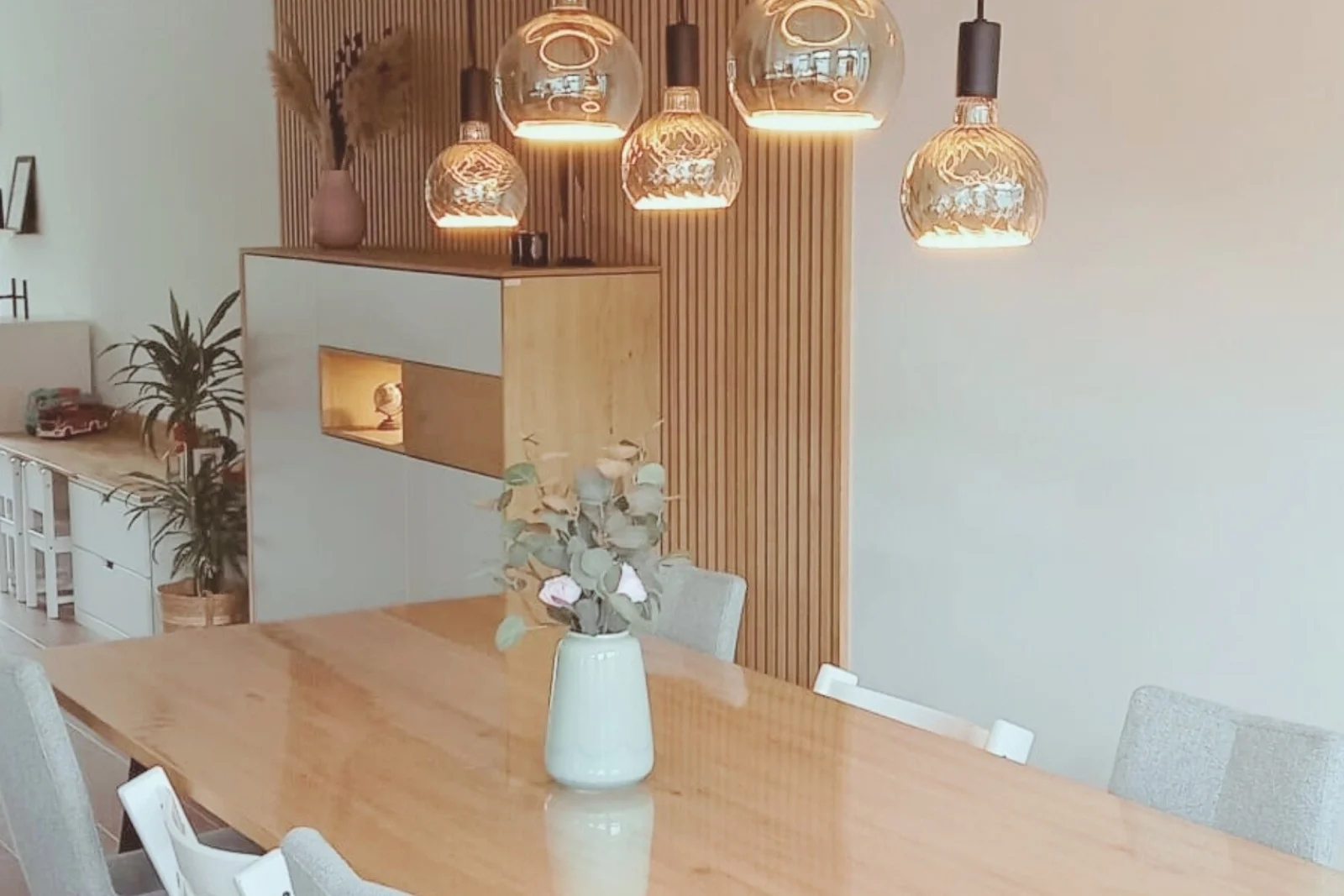 Moderne eethoek met houten eettafel, vaas met bloemen en glazen hanglampen, met een wand van houten latten en een kast met nisverlichting op de achtergrond.