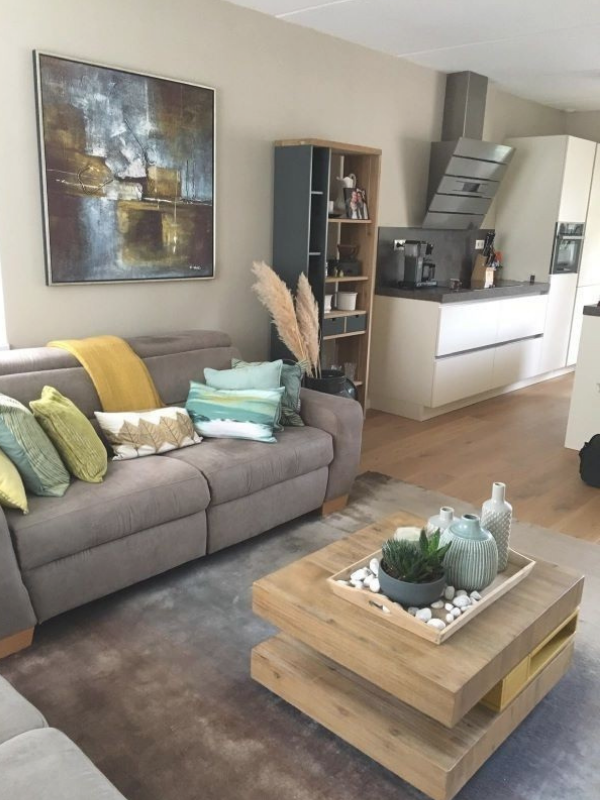 Deze foto toont het totaalbeeld van de woonkamer & eetkamer make-over, met een warme, rustige basis en een fijne open verbinding met de keuken. De taupe bank en het zachte vloerkleed zorgen voor comfort, terwijl het houten salontafelblad warmte en na