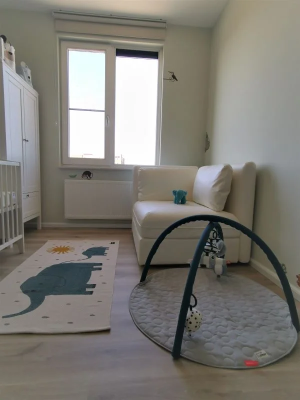 Rustige, lichte babykamer met zachte groentint op de muren en een houtlook vloer. Links staan een wit ledikant en een hoge witte kast. Het grote raam met rolgordijn brengt veel daglicht binnen, met een radiator eronder. Rechts staat een compact wit b