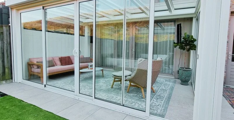 Glazen veranda/serre met schuifwanden, houten loungebank, relaxstoel en groot vloerkleed.