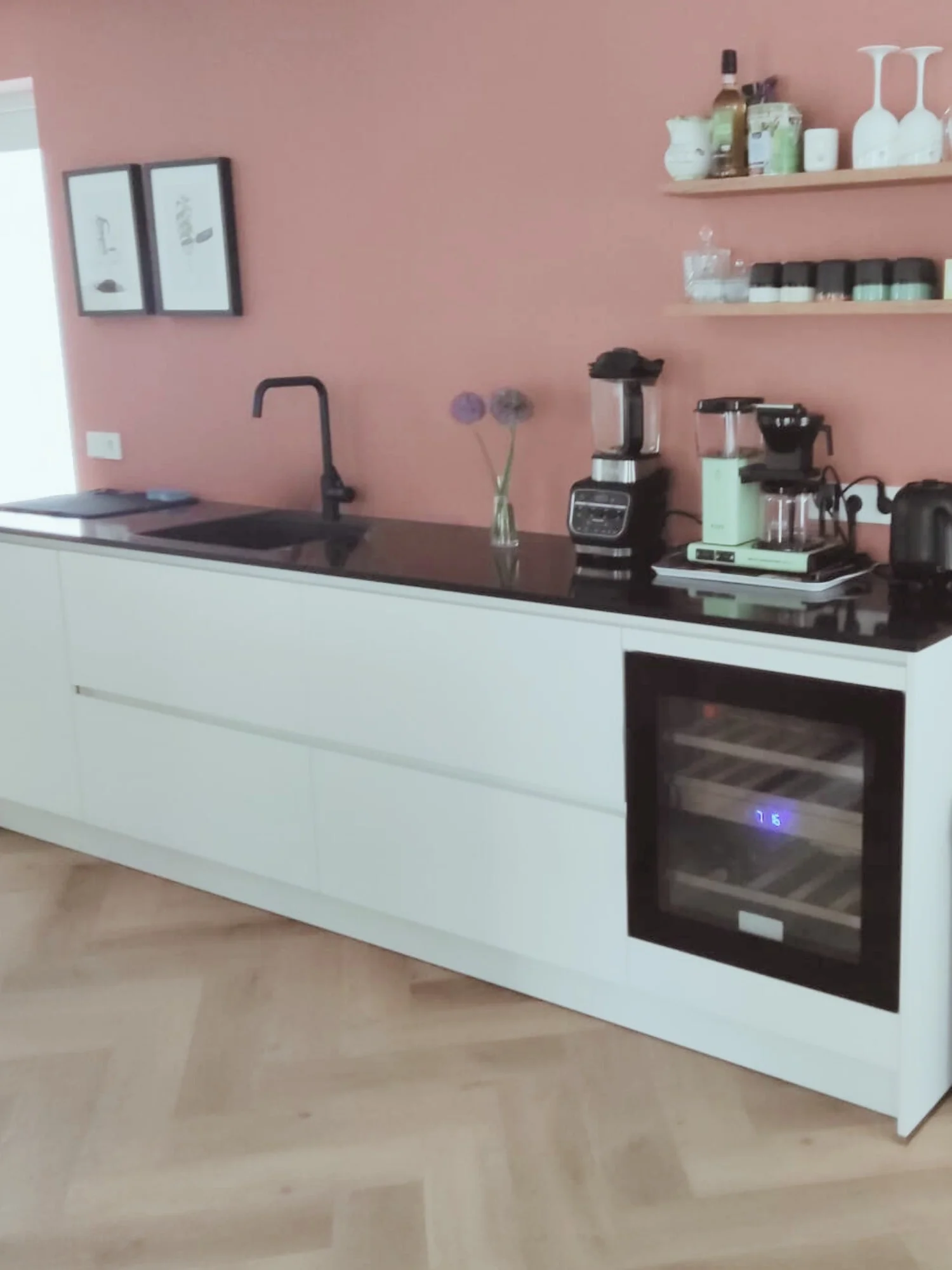 Strakke keukenwand met witte, greeploze onderkasten en een zwart werkblad. Tegen de zachtroze muur staan een zwarte kraan en spoelbak, een ingebouwde wijnkoelkast en diverse keukenapparaten. Open houten planken met glazen, kruiden en accessoires zorg