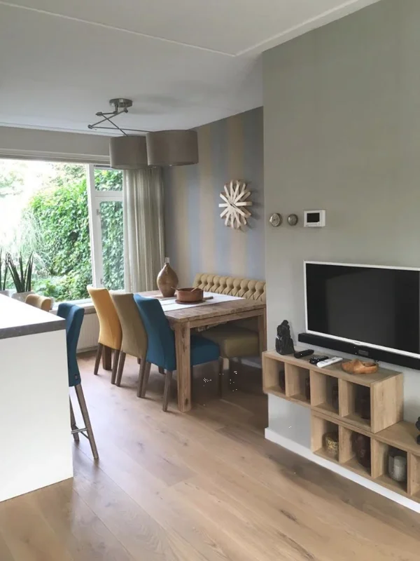 Deze foto laat het totaalbeeld zien van de woonkamer & eetkamer make-over, met een open indeling, veel daglicht en een warme, rustige basis. De houten vloer en het natuurlijke tv-meubel brengen warmte, terwijl de groene accentmuur zorgt voor sfeer en