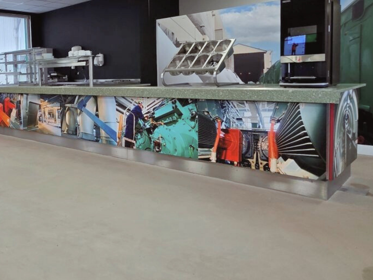 Detail van de toonbank met een fotoprint die het verhaal van de fabriek door de jaren heen laat zien. De beelden van machines, werkzaamheden en industriële onderdelen geven de ruimte een stoer en herkenbaar karakter, terwijl de strakke afwerking zorg