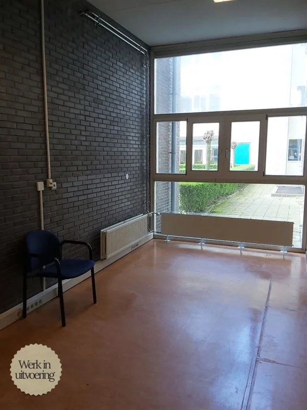 Lege docentenstudiekamer klaar voor nieuwe inrichting, met industriële bakstenen muur, hoge plafonds en grote ramen met veel daglicht als basis voor een moderne studie- en werkplek.
