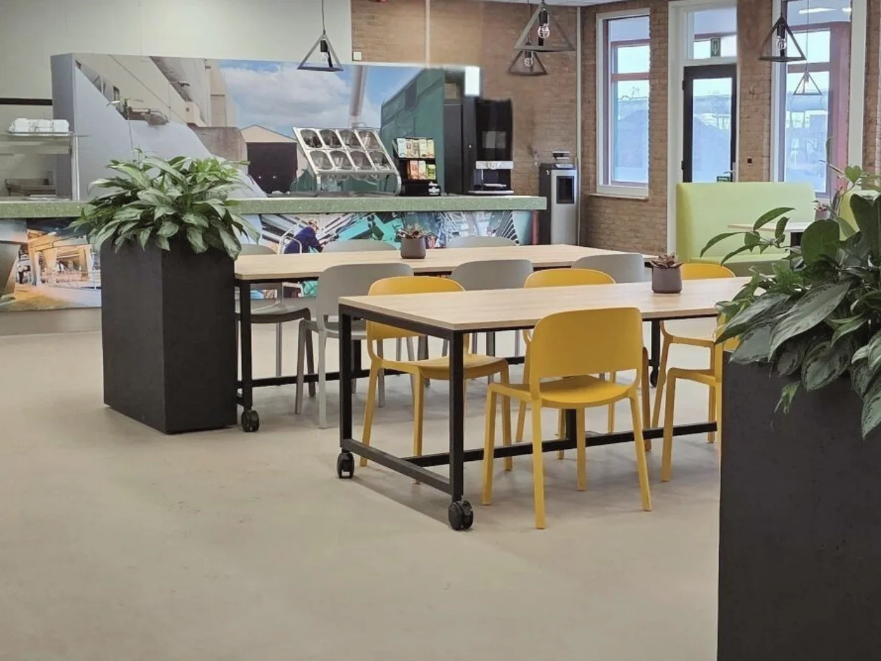 Een lichte, uitnodigende zithoek in de kantine met verrijdbare tafels en warme gele stoelen, omringd door grote planten voor een frisse, groene touch. Op de achtergrond geeft de lange toonbank met fotoprint van de fabriek een stoer, industrieel accen
