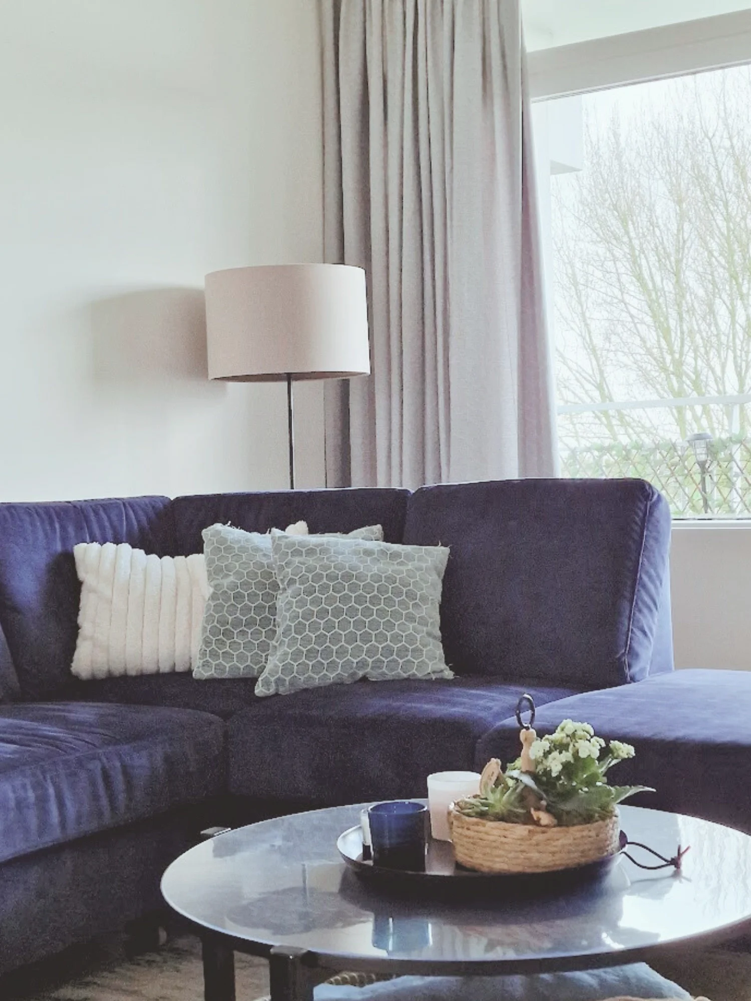 Close-up van een gezellige zithoek met donkerblauwe bank, zachte kussens en ronde salontafel. Neutrale tinten en lichtinval geven rust.  Interieuradvies, interieurontwerp en styling door En-Casa Interiors in Noord-Holland.