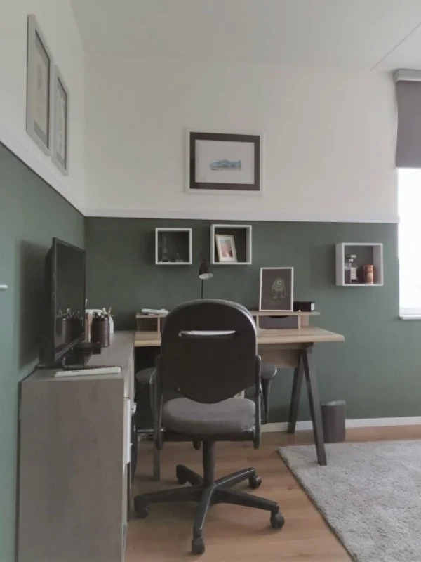 Deze werkplek is stoer en functioneel tegelijk. En-Casa combineerde warm hout met zwart accent en groen behang in de lambrisering, zodat alles één geheel vormt met de rest van de kamer. De wandkubussen zorgen voor persoonlijke styling zonder dat het 