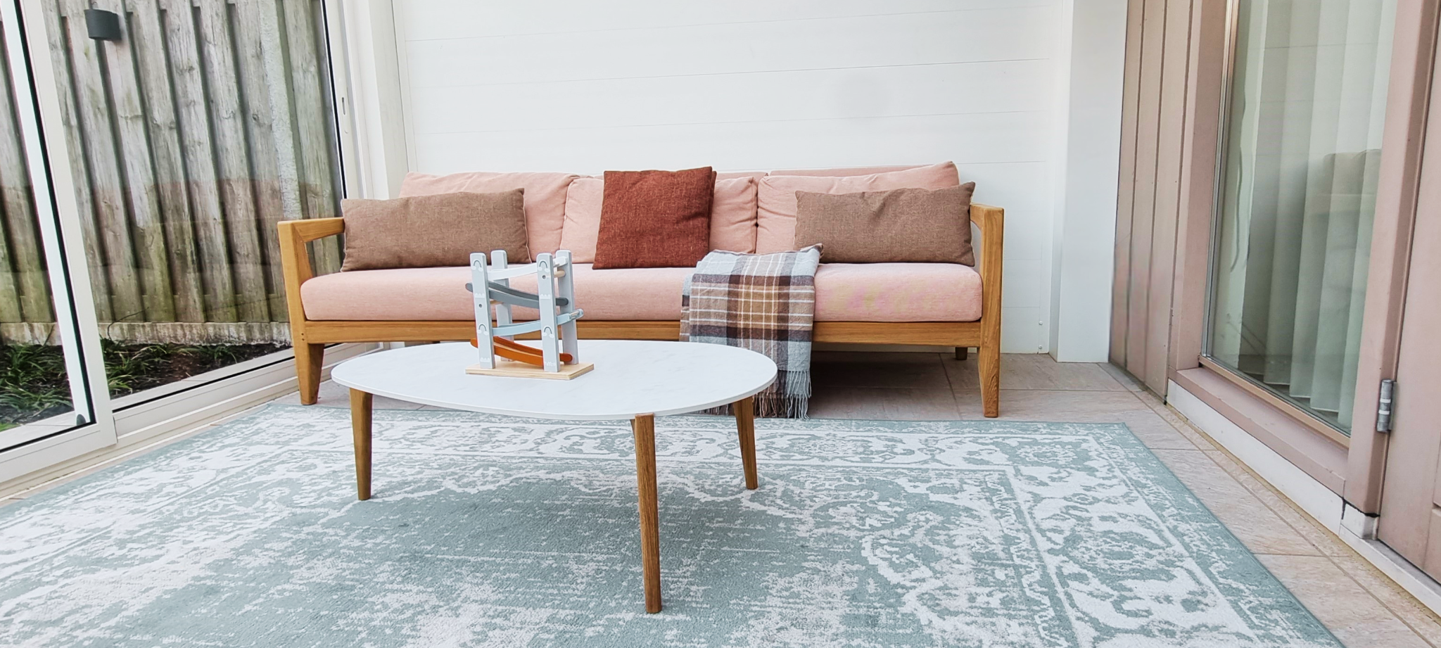 Veranda zithoek met houten bank, kussens in roze en terracotta, ronde salontafel en een licht patroonvloerkleed.