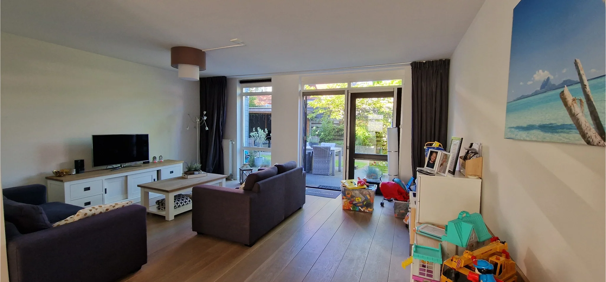 Voorfoto van een lichte woonkamer met zithoek, tv-meubel en grote schuifpui naar de tuin, met speelplek voor kinderen.
