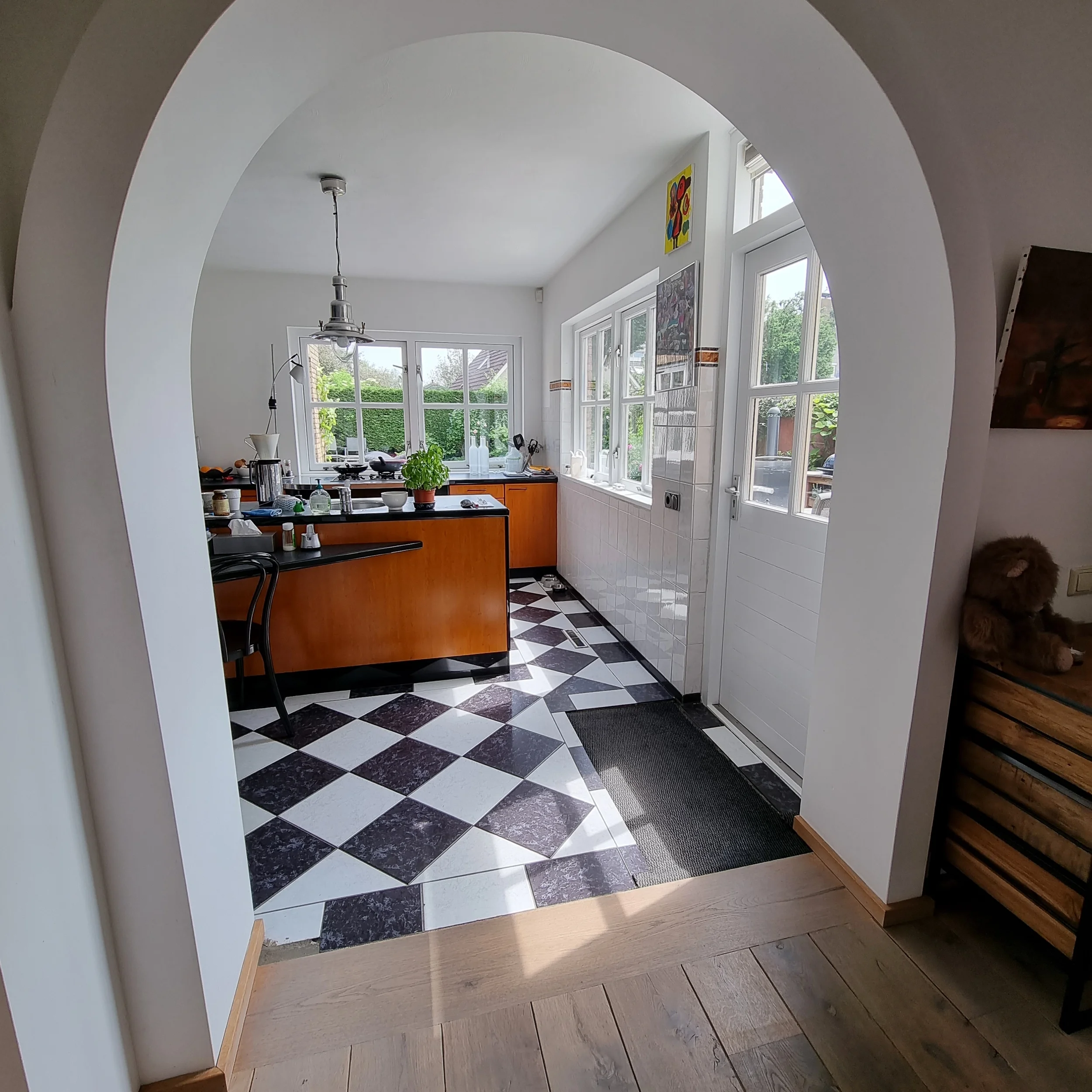 Before foto met doorkijk naar de keuken door een ronde boogdoorgang, met zwart-witte tegelvloer, houten keukenkasten en veel daglicht. De ruimte oogt functioneel en licht, maar mist nog een rustige eenheid en samenhang met de rest van de begane grond
