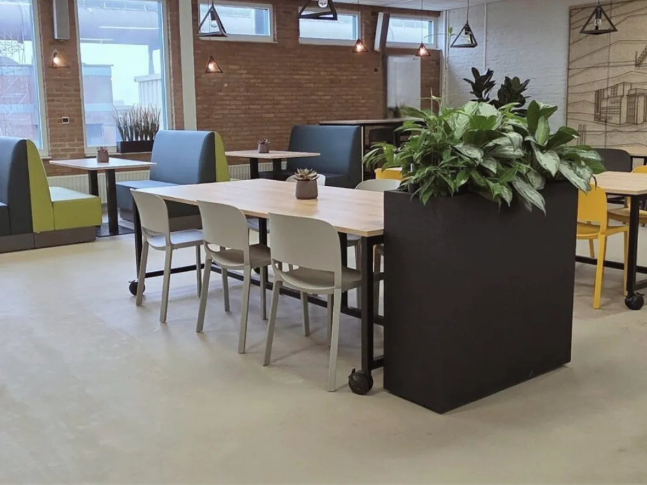 Een rustige werk- en zitplek in de kantine met een lange tafel, comfortabele stoelen en praktische schermen voor extra focus en privacy. De neutrale vloer en het warme baksteen, gecombineerd met het zachte daglicht uit de hoge ramen, geven de ruimte 