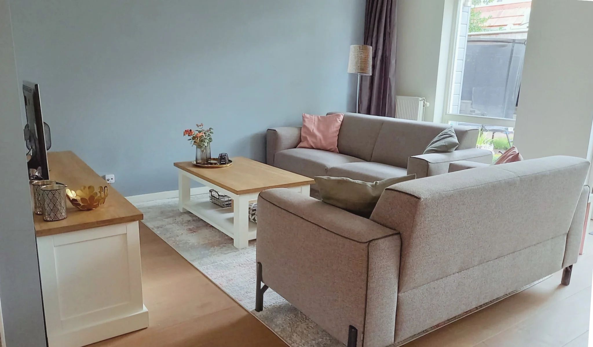 Gezellige zithoek met grijze bank, zachte pastel kussens en een lichte houten salontafel. Rustige basis met warm daglicht en natuurlijke materialen voor een comfortabele, moderne en casa.  Interieuradvies, interieurontwerp en styling door En-Casa Int
