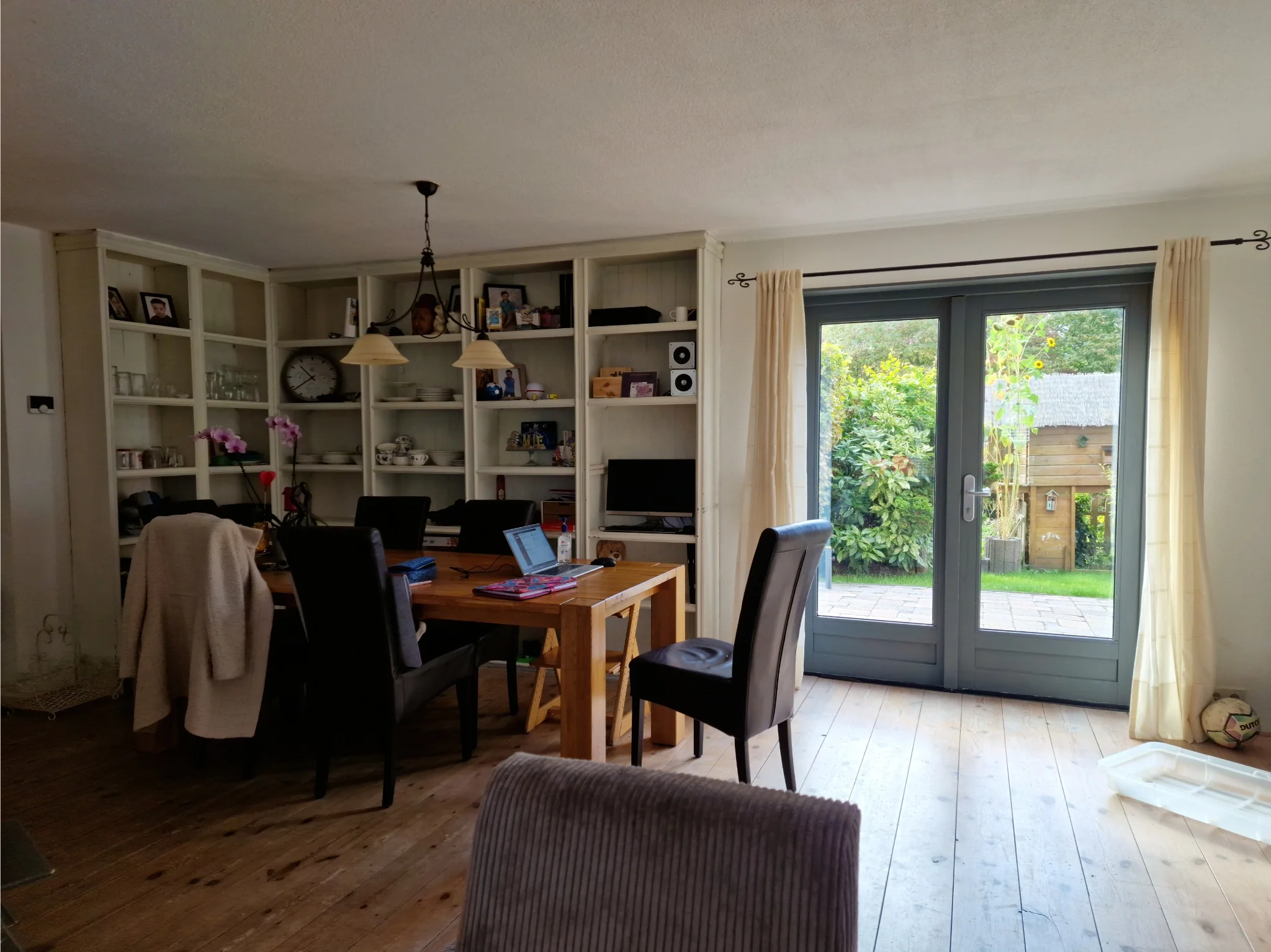 Eethoek in before-situatie met houten eettafel en zwarte stoelen, ingebouwde open kastenwand met servies en persoonlijke items, en dubbele tuindeuren.