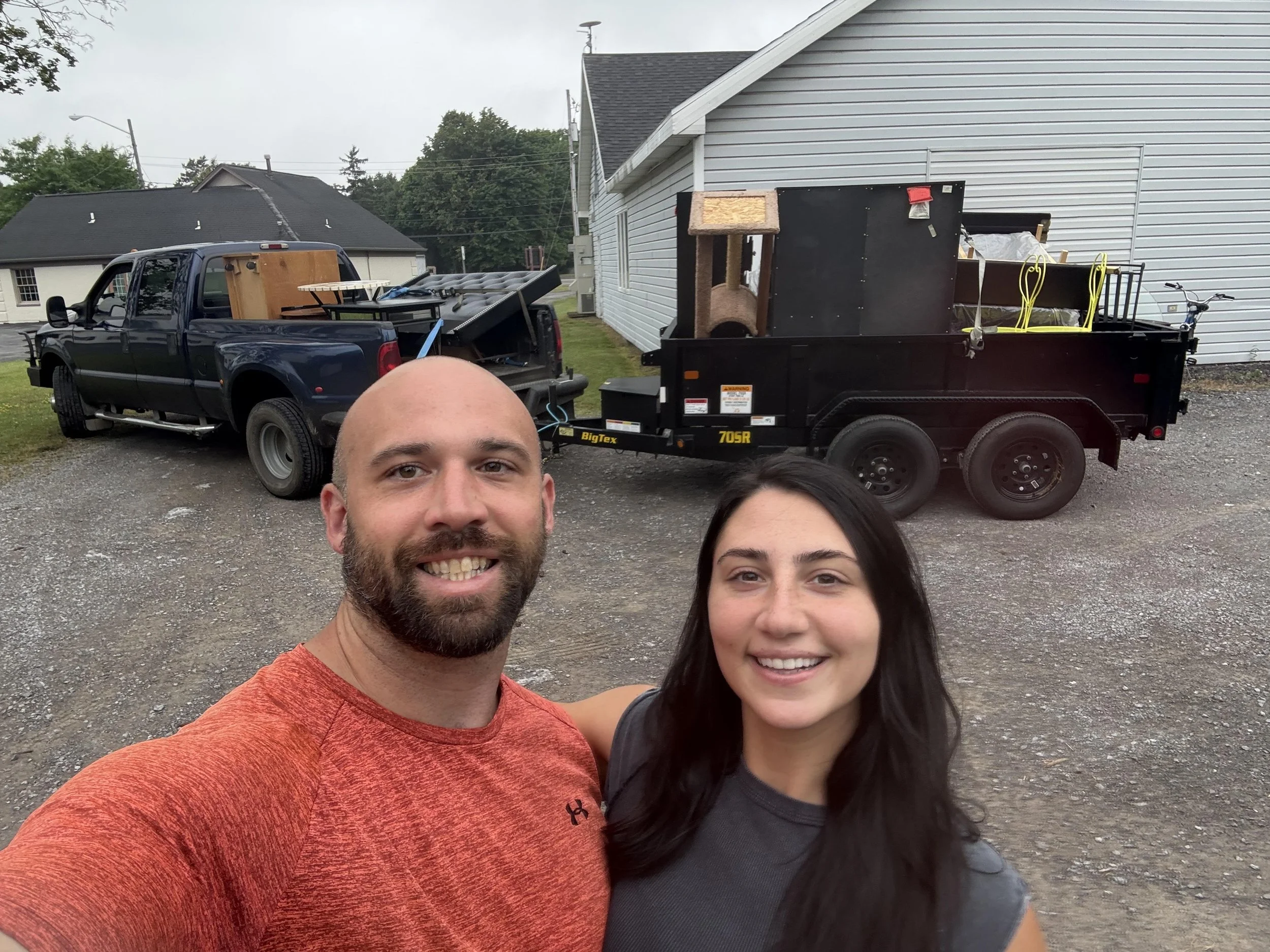 A man and woman smiling for a selfie outdoors, with trucks and a trailer loaded with furniture and belongings in the background.