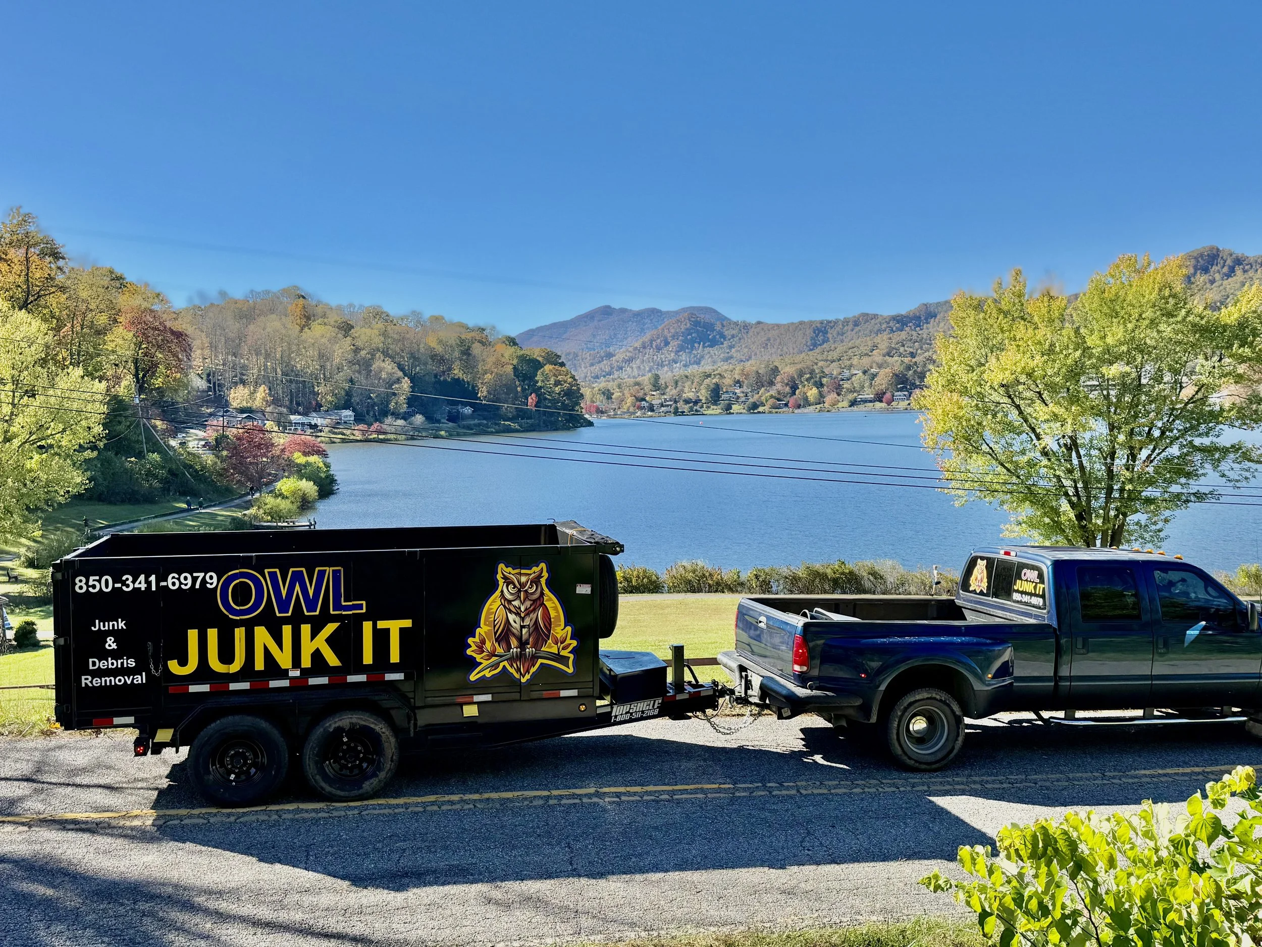 A black junk removal truck labeled 'OWL JUNK IT' with a cartoon owl logo, attached to a black pickup truck, parked on a road overlooking a lake with trees and mountains in the background.