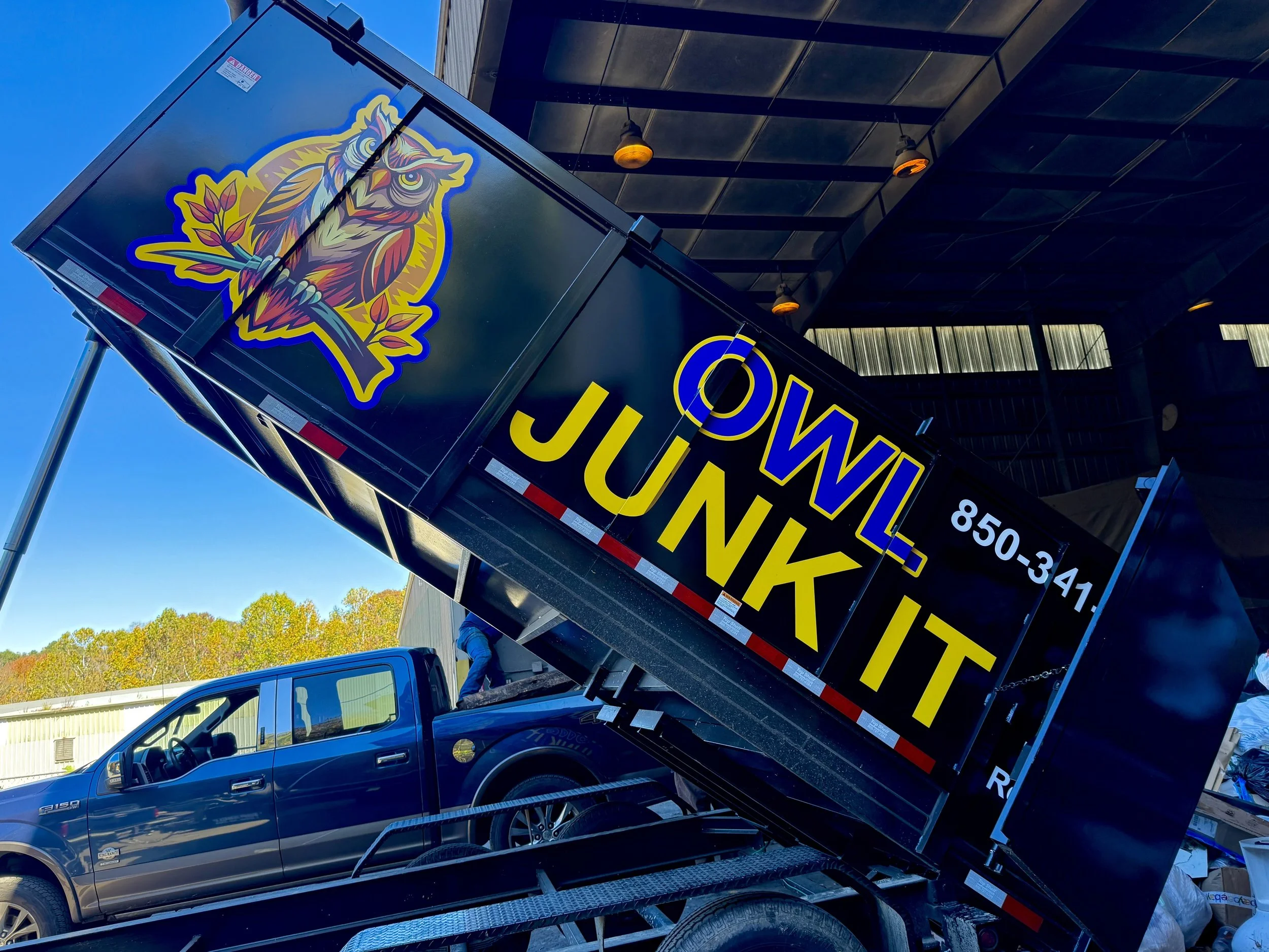 A black moving truck with a colorful owl logo and the words "OWL JUNKIT" in large yellow letters, loaded with items, parked in front of a building under a covered area with orange lights.