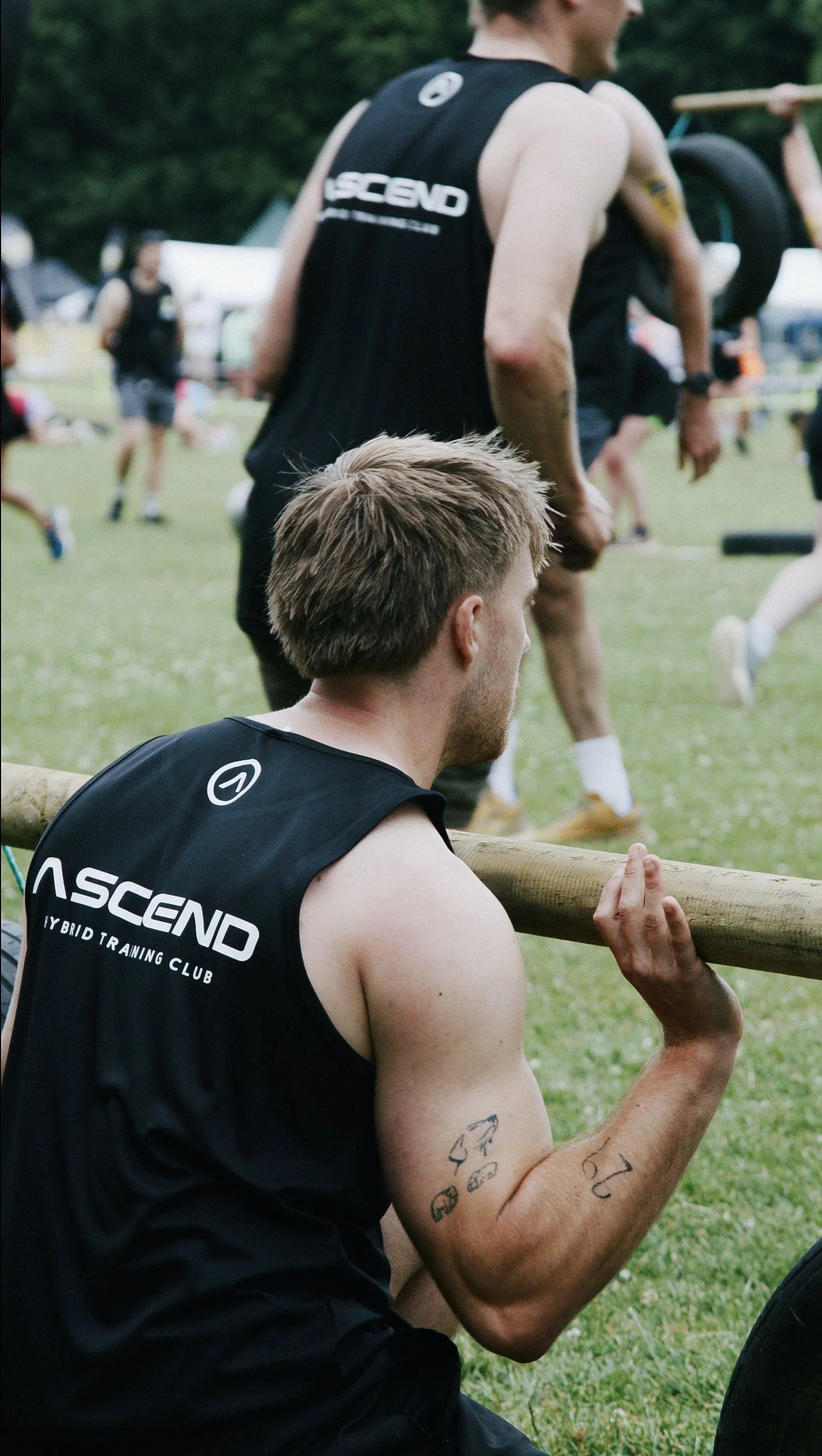 A man with tattoos on his arm wearing a black sleeveless athletic shirt, sitting on the grass and holding a wooden log across his shoulders during a fitness event, with other people in athletic attire and tents in the background.