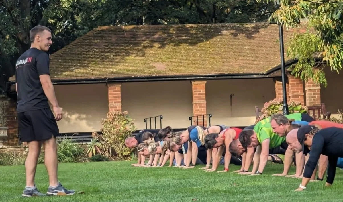 A group of people performing push-ups outdoors on a grassy lawn, with a man standing on the side smiling and observing.