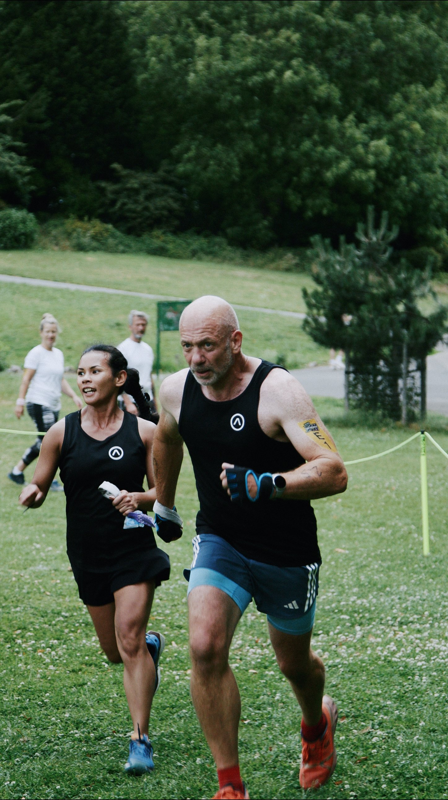A group of people running outdoors in a grassy park area with trees in the background. The focus is on a man and a woman in the front, both wearing black athletic clothing, participating in a race or running event.