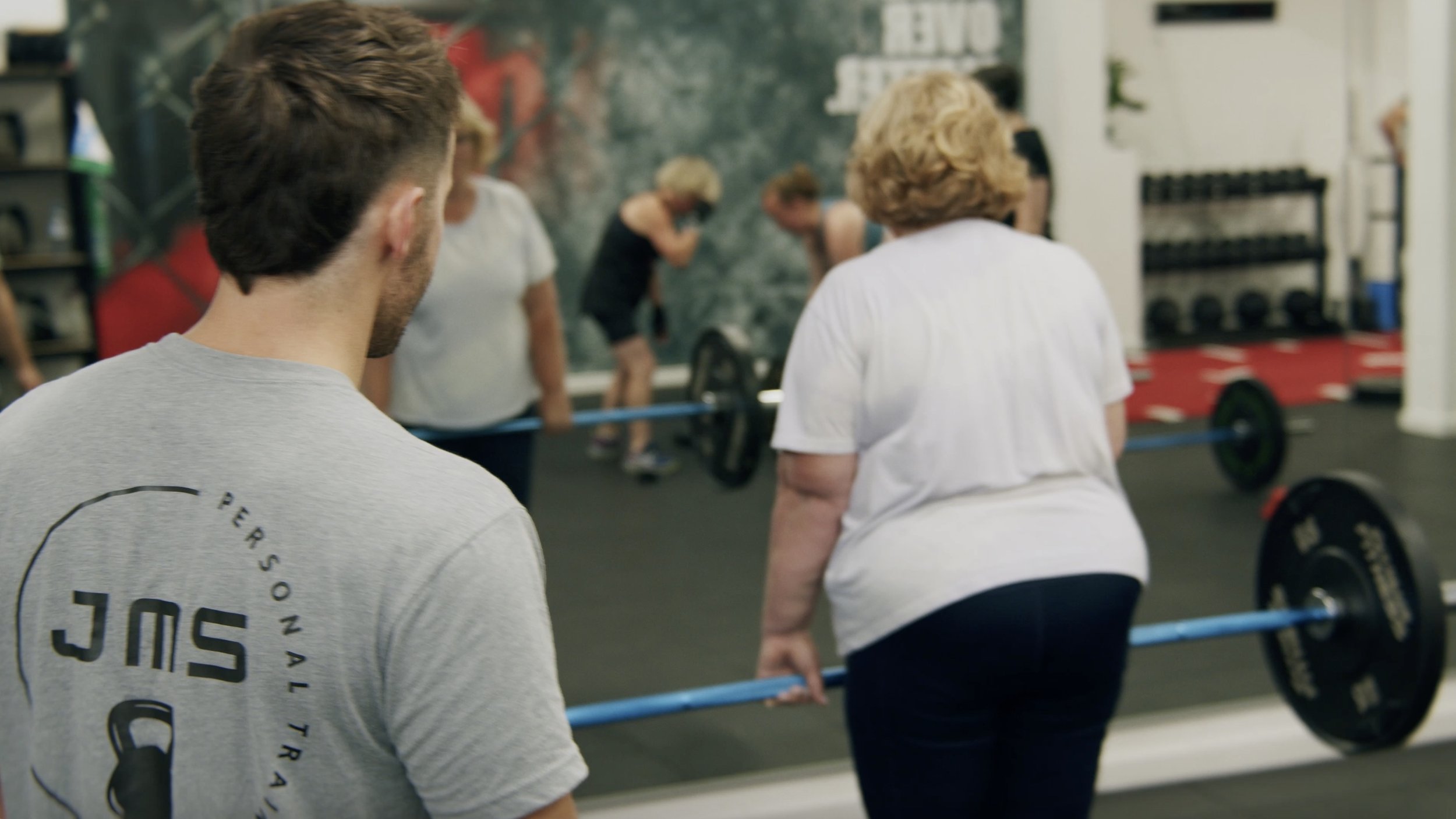 People lifting weights at a gym, with a woman in a white shirt lifting a barbell while others watch.
