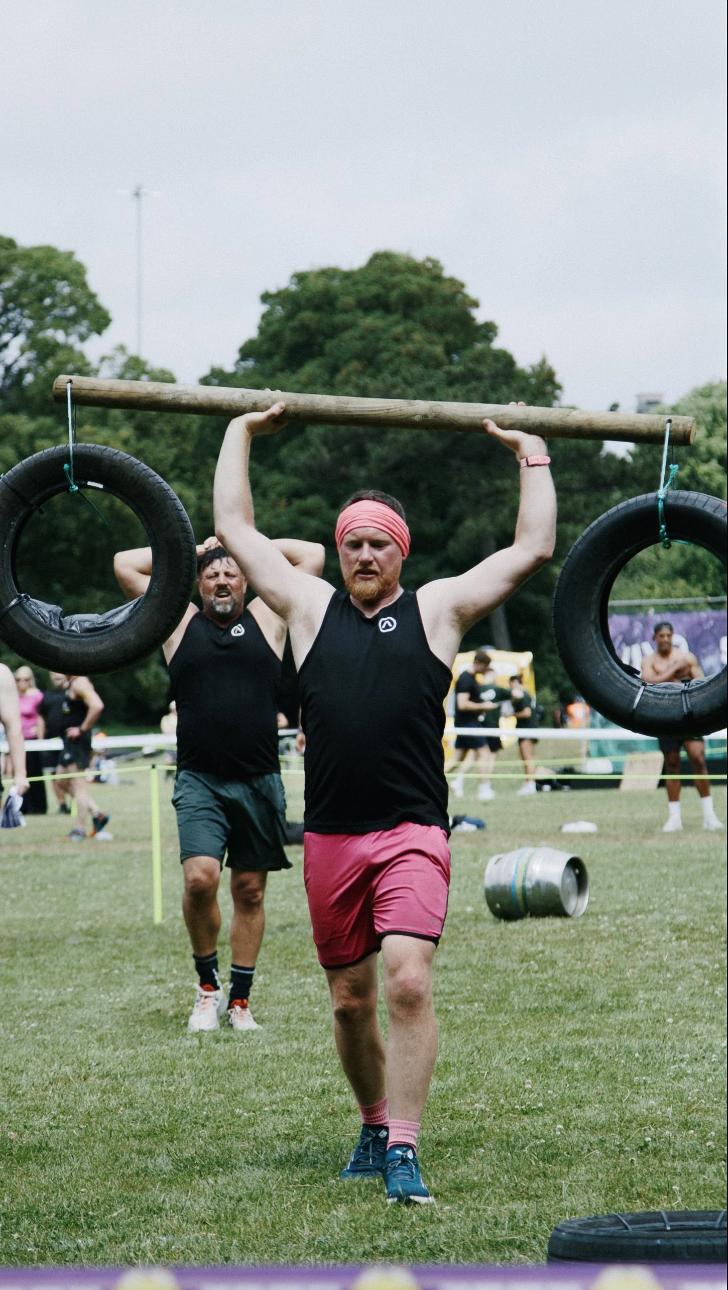 A man with a red headband and beard carrying tires on a wooden pole during an outdoor fitness competition, with a grassy field and other people in the background.