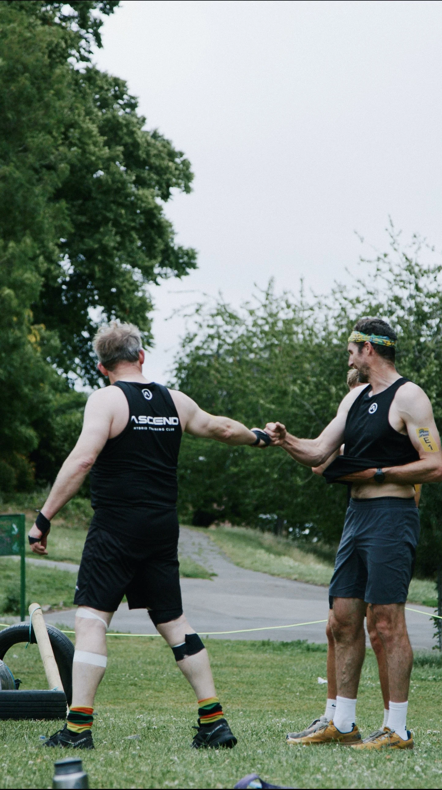 Two men in athletic clothing shake hands outdoors, surrounded by trees and grass, after a fitness event.