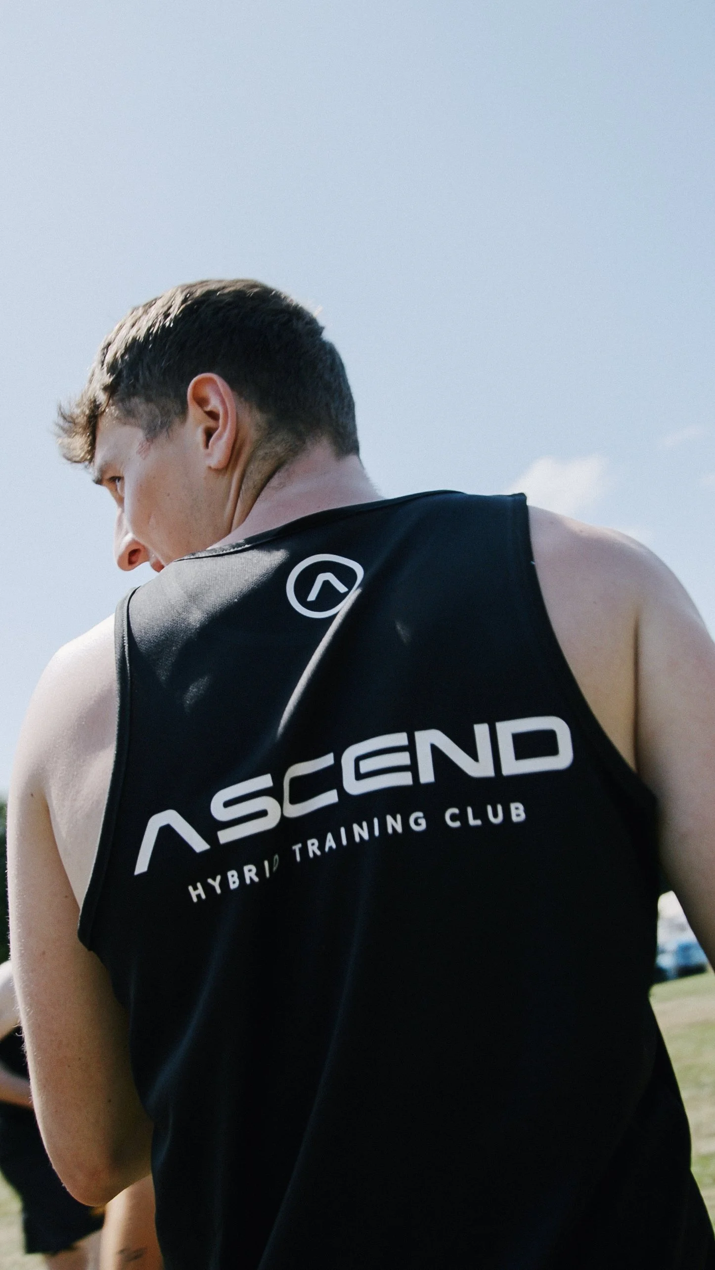 Young male athlete in black sleeveless shirt with "ASCEND HYBRID TRAINING CLUB" logo on back, outdoors on sunny day.