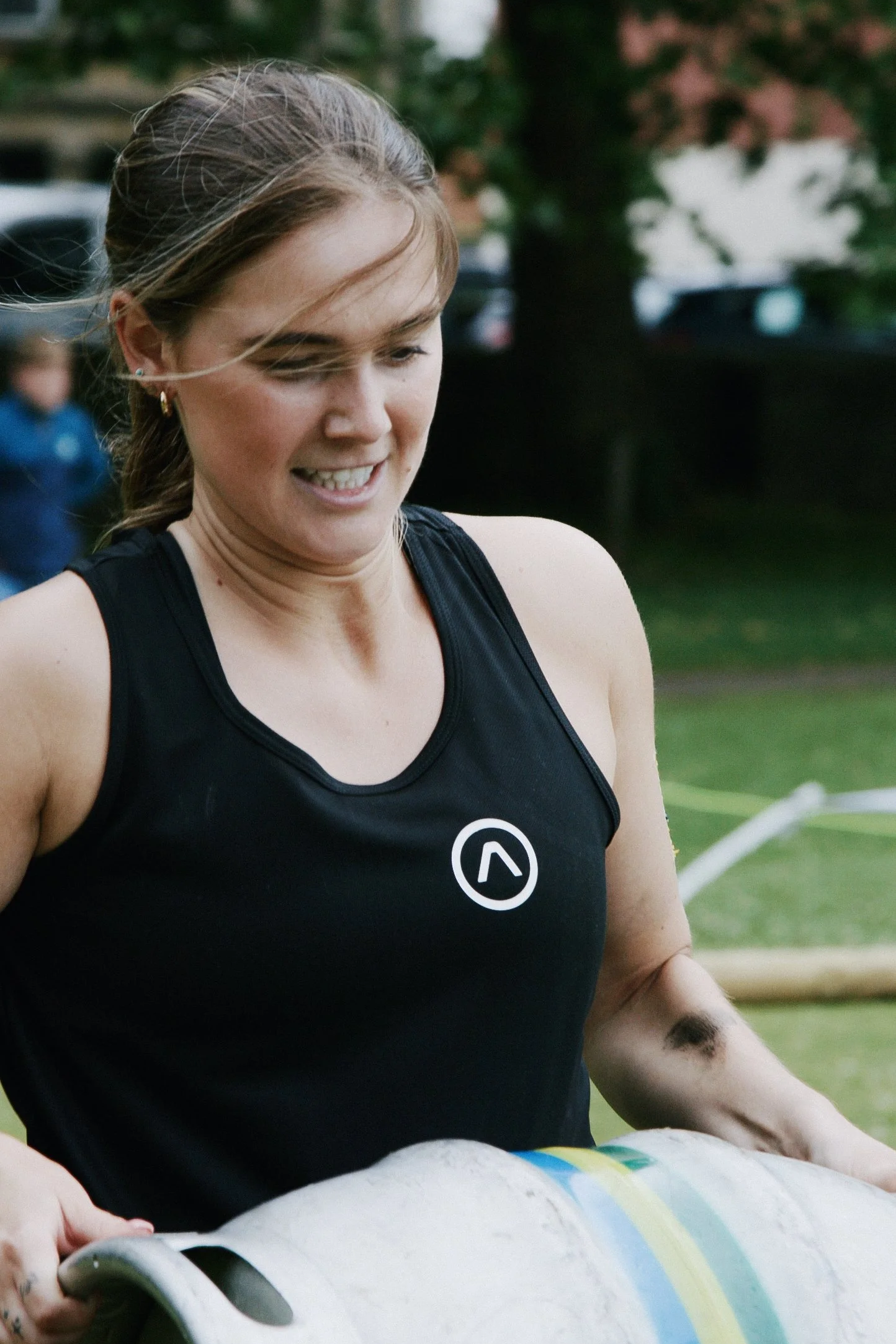 A woman in a black athletic tank top holding a rolled-up sleeping mat outdoors, smiling with a participate in an event or race.