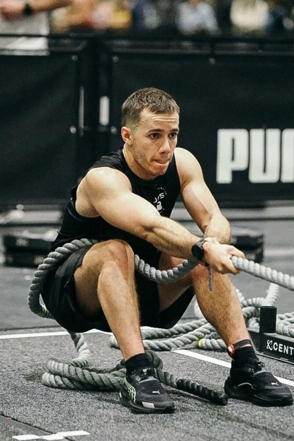 Man with short hair and tattooed arms doing tire-flip exercise with battle ropes at gym.