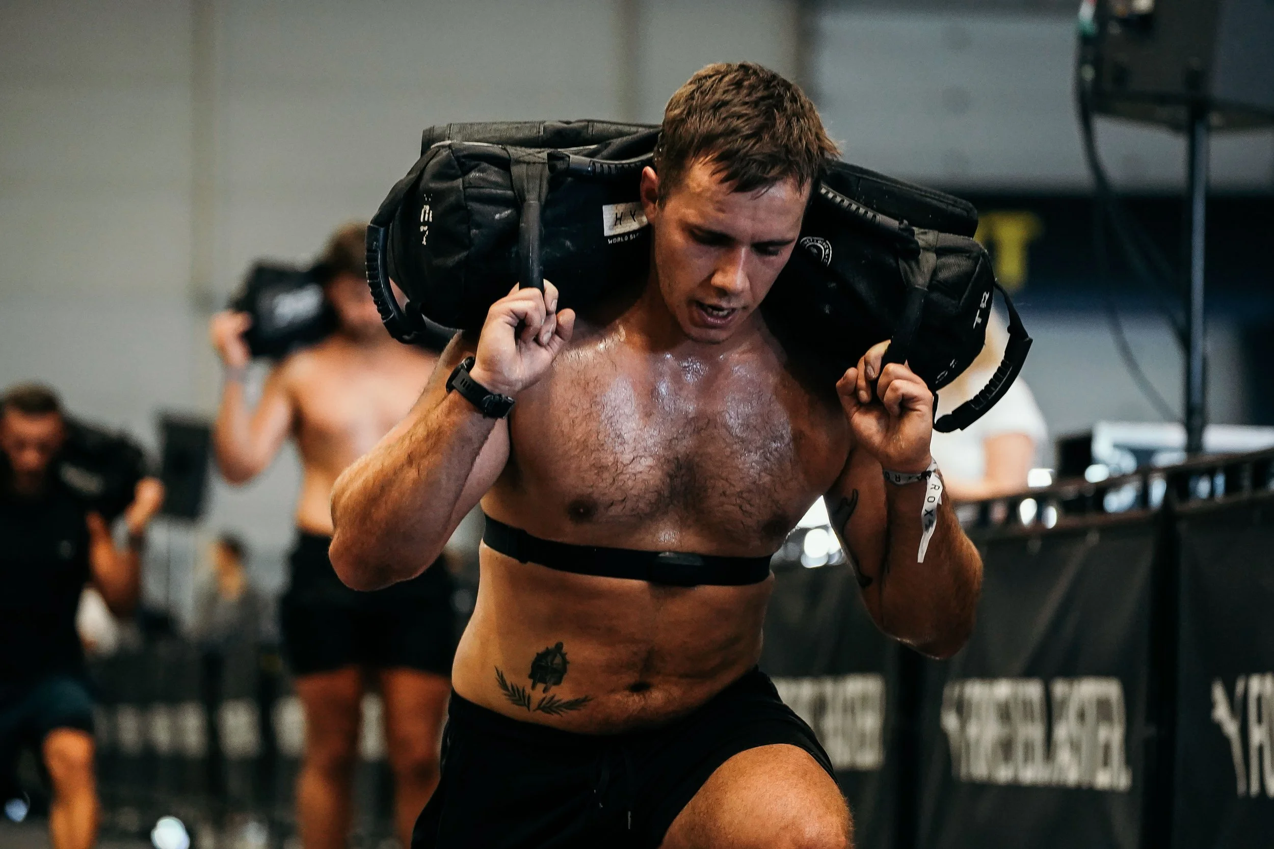 A shirtless man carrying a weighted pack on his shoulders during a workout in a gym.