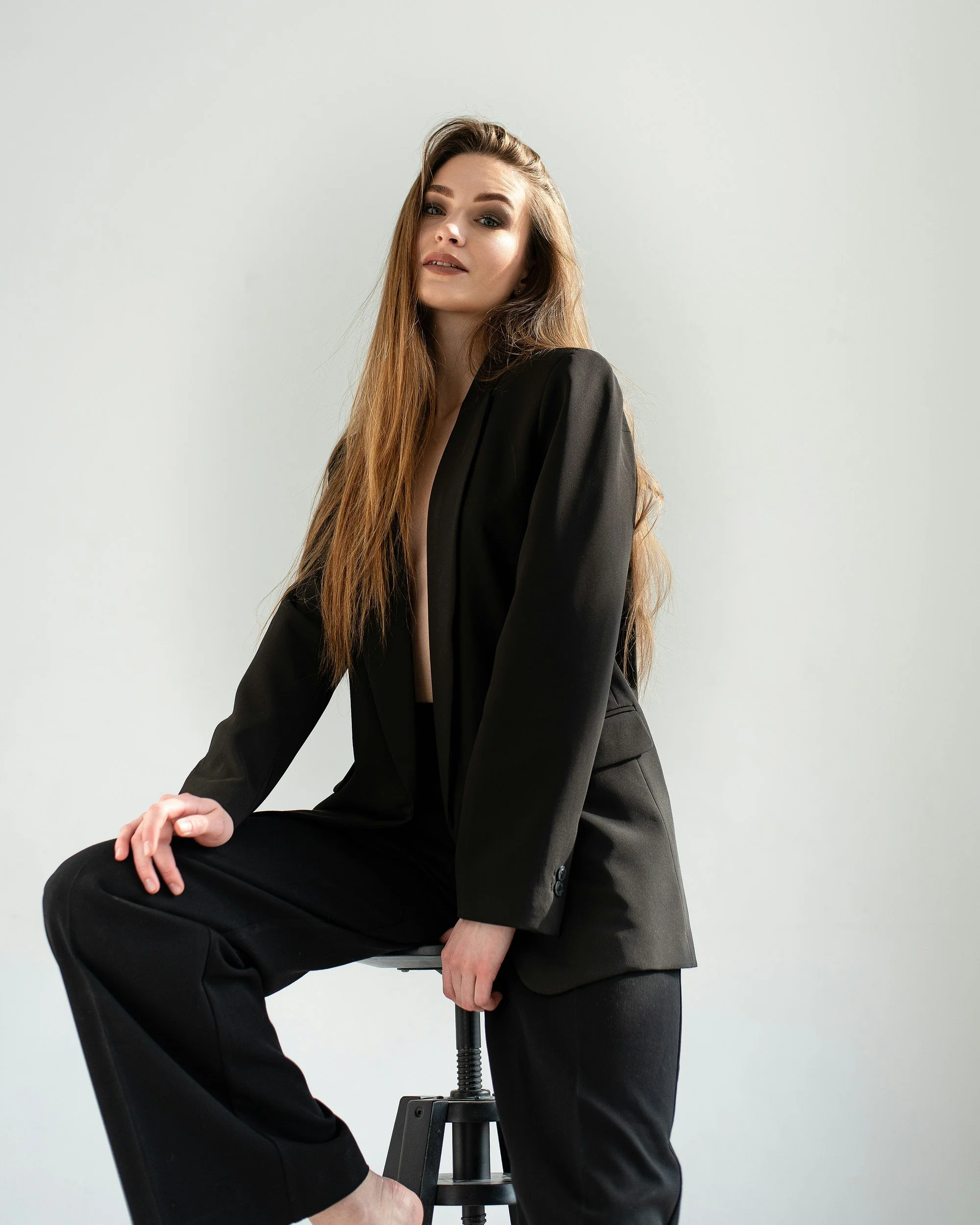 Femme en tailleur noir posant sur un tabouret contre un mur blanc