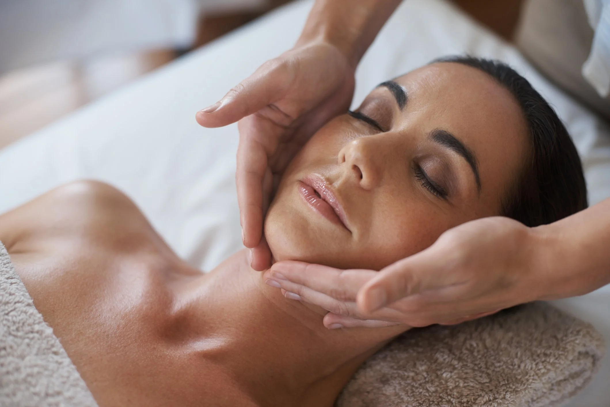 A woman receiving a facial massage from a therapist, with her eyes closed and a relaxed expression.
