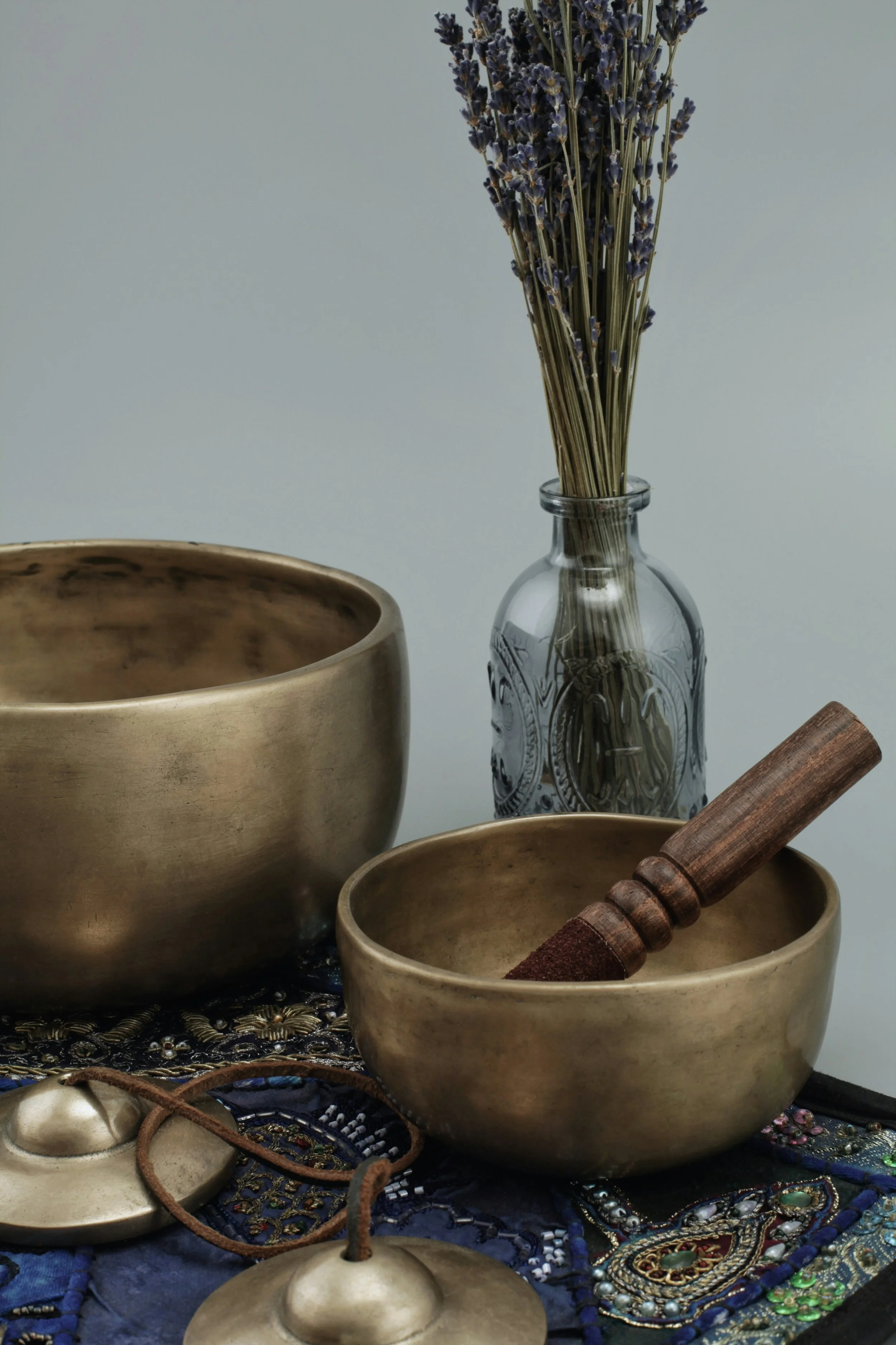 Two large brass bowls, a wooden mallet, and small brass cymbals on an embroidered dark blue cloth, with lavender flowers in a glass bottle in the background.