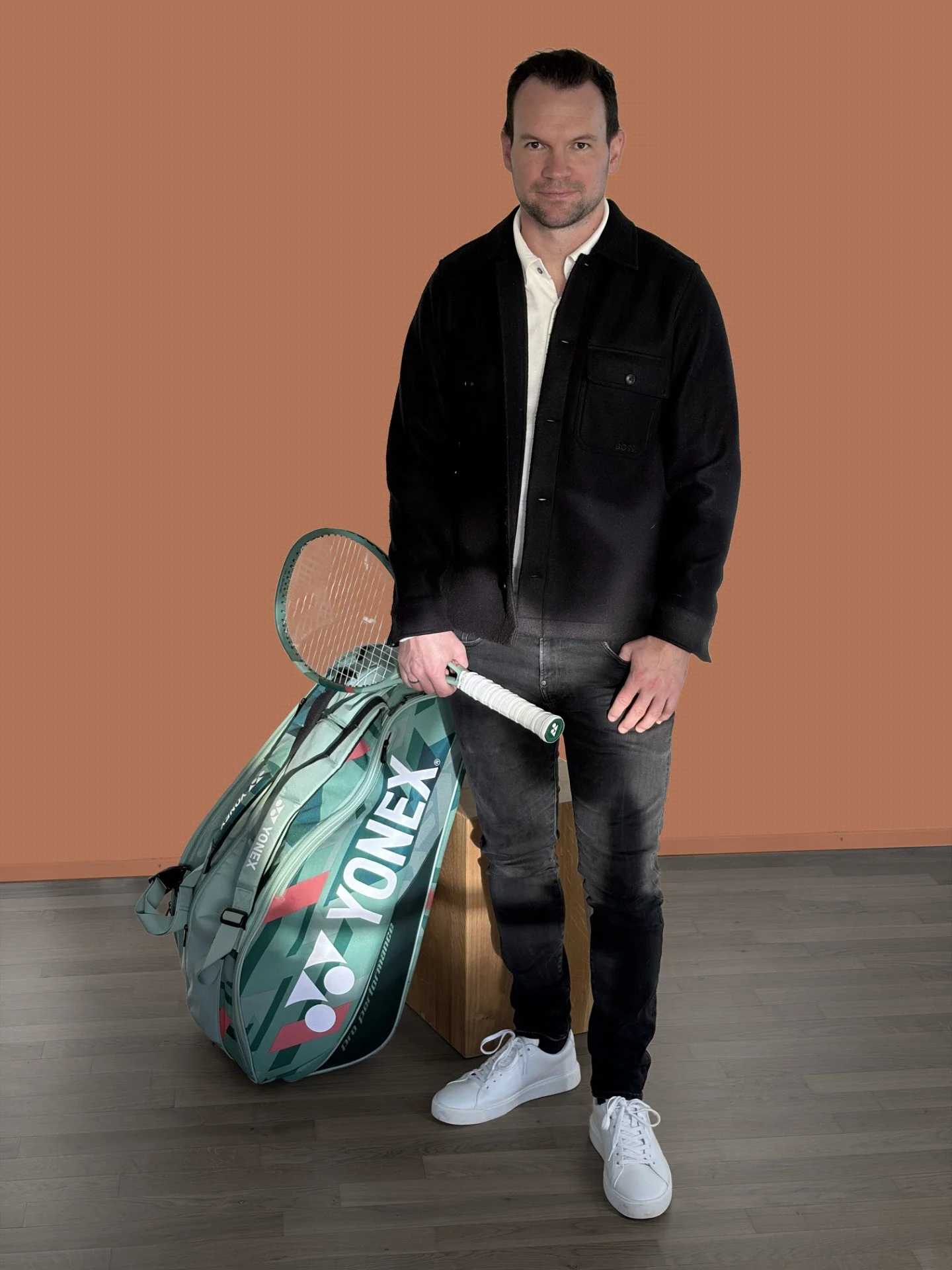 Mann steht mit Tennisschläger und Tennistasche vor einem braunen Hintergrund, trägt weiße Schuhe, schwarze Jeans und eine schwarze Jacke.