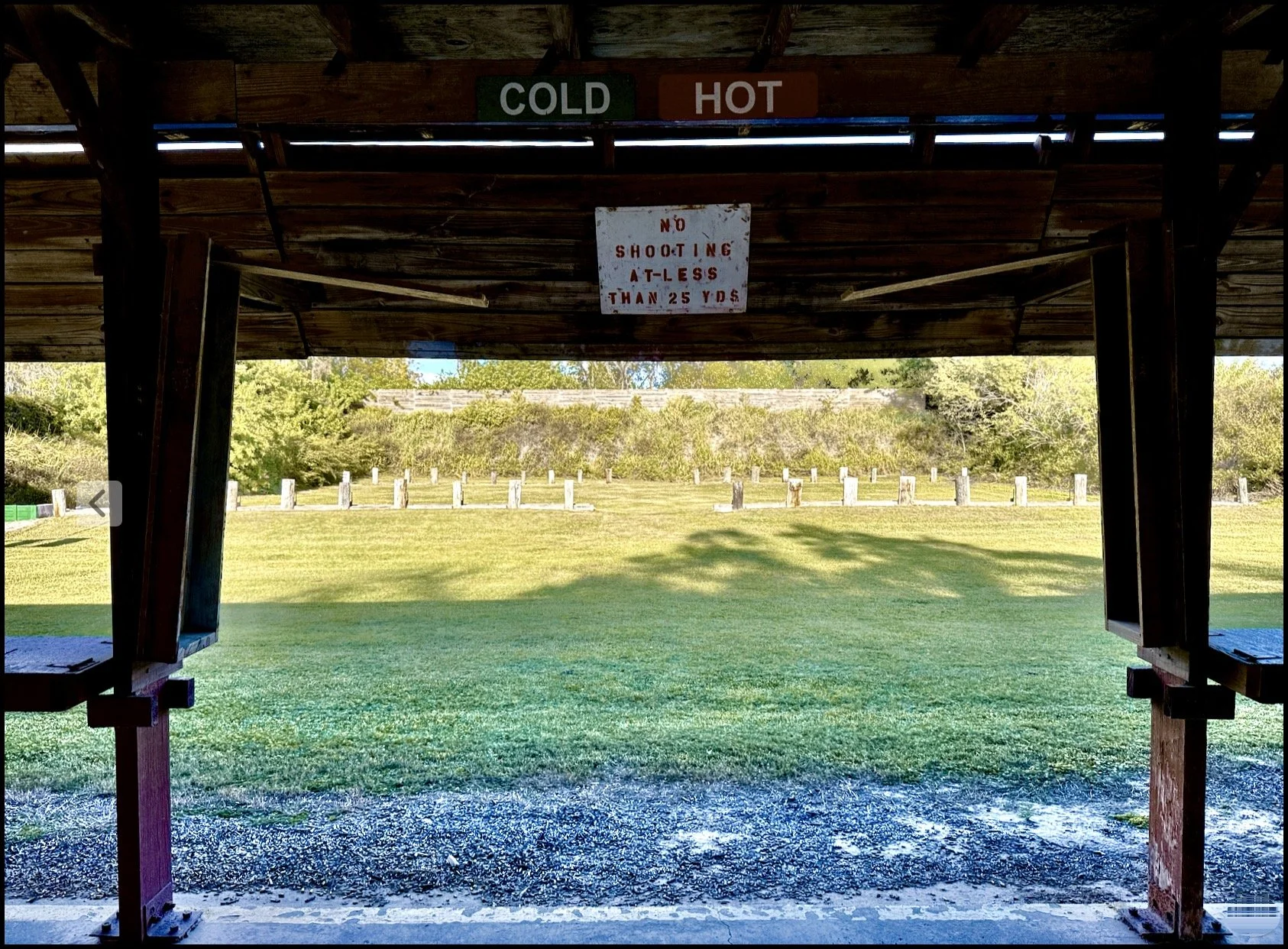 View from under a wooden shelter with a sign that reads 'No Shooting At Less Than 25 Yards' outdoors with grass, trees, and a row of wooden posts.
