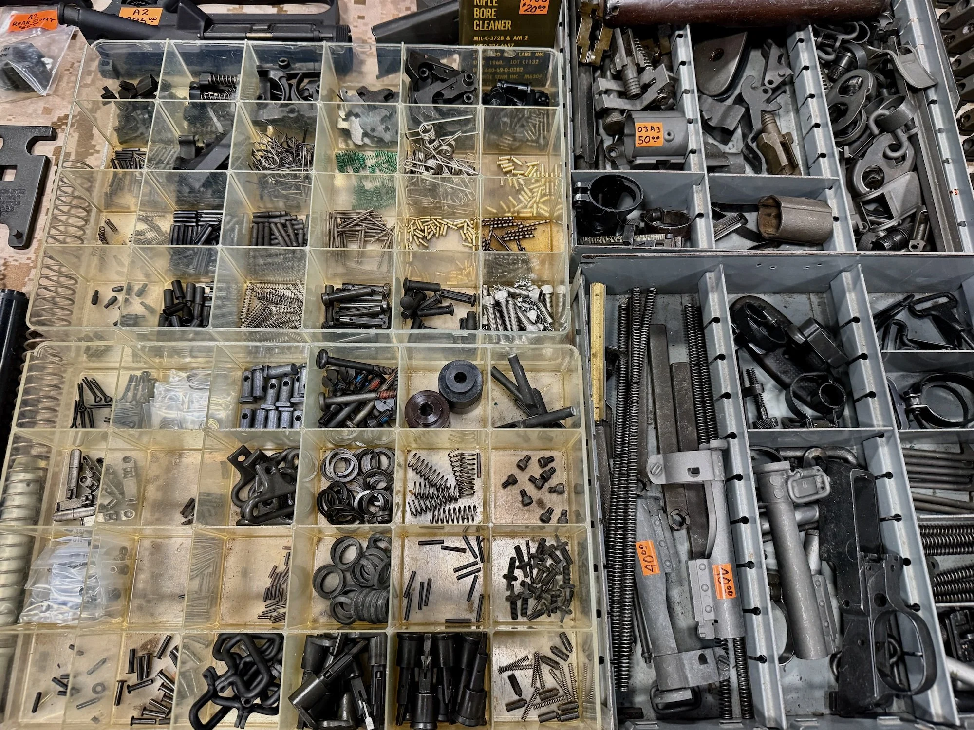 Assorted firearm parts, including small metal fasteners, springs, screws, and mechanical parts organized in clear plastic compartment trays.